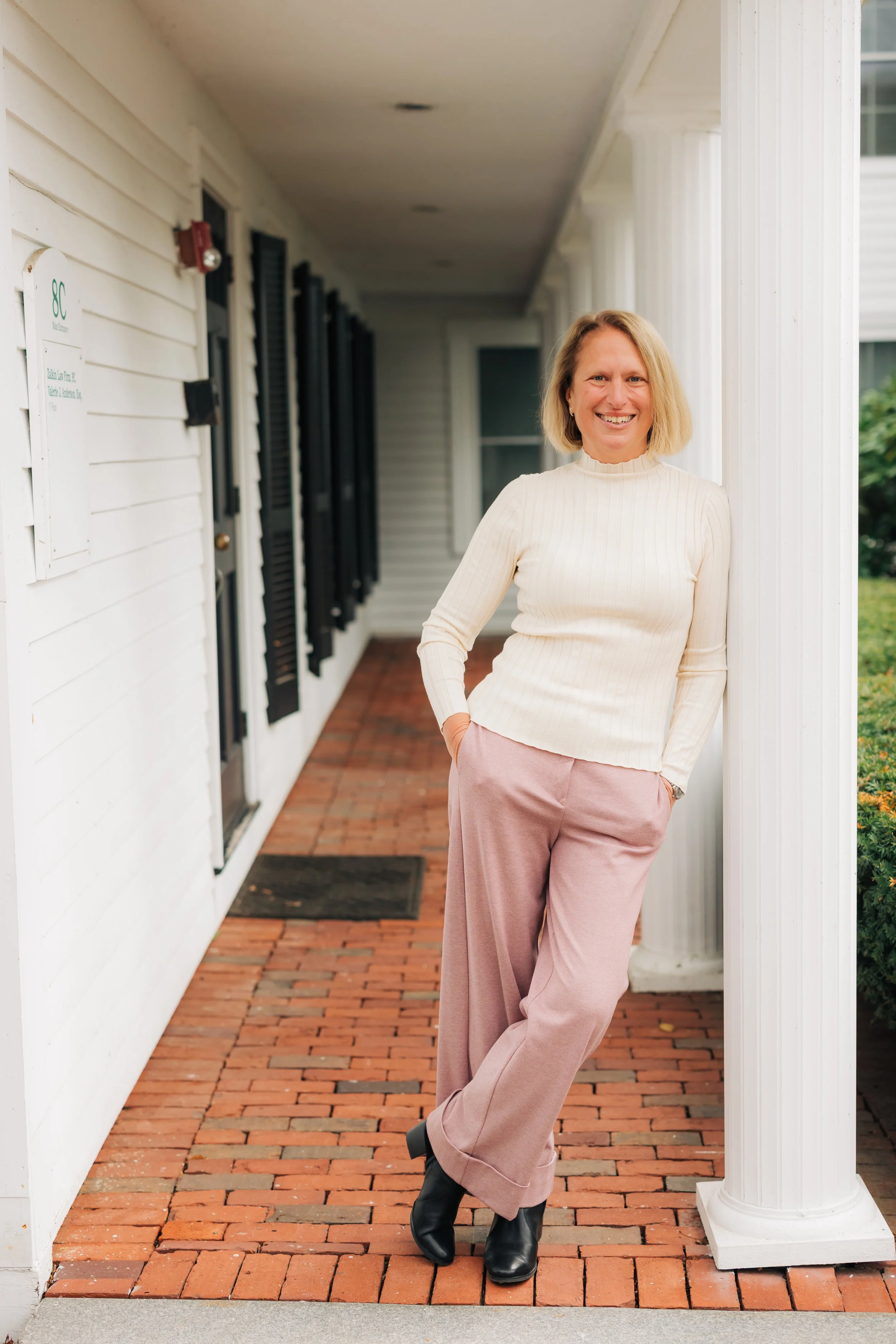 A woman with blonde hair smiling standing on a brick sidewalk outside a building with white siding and black shutters.