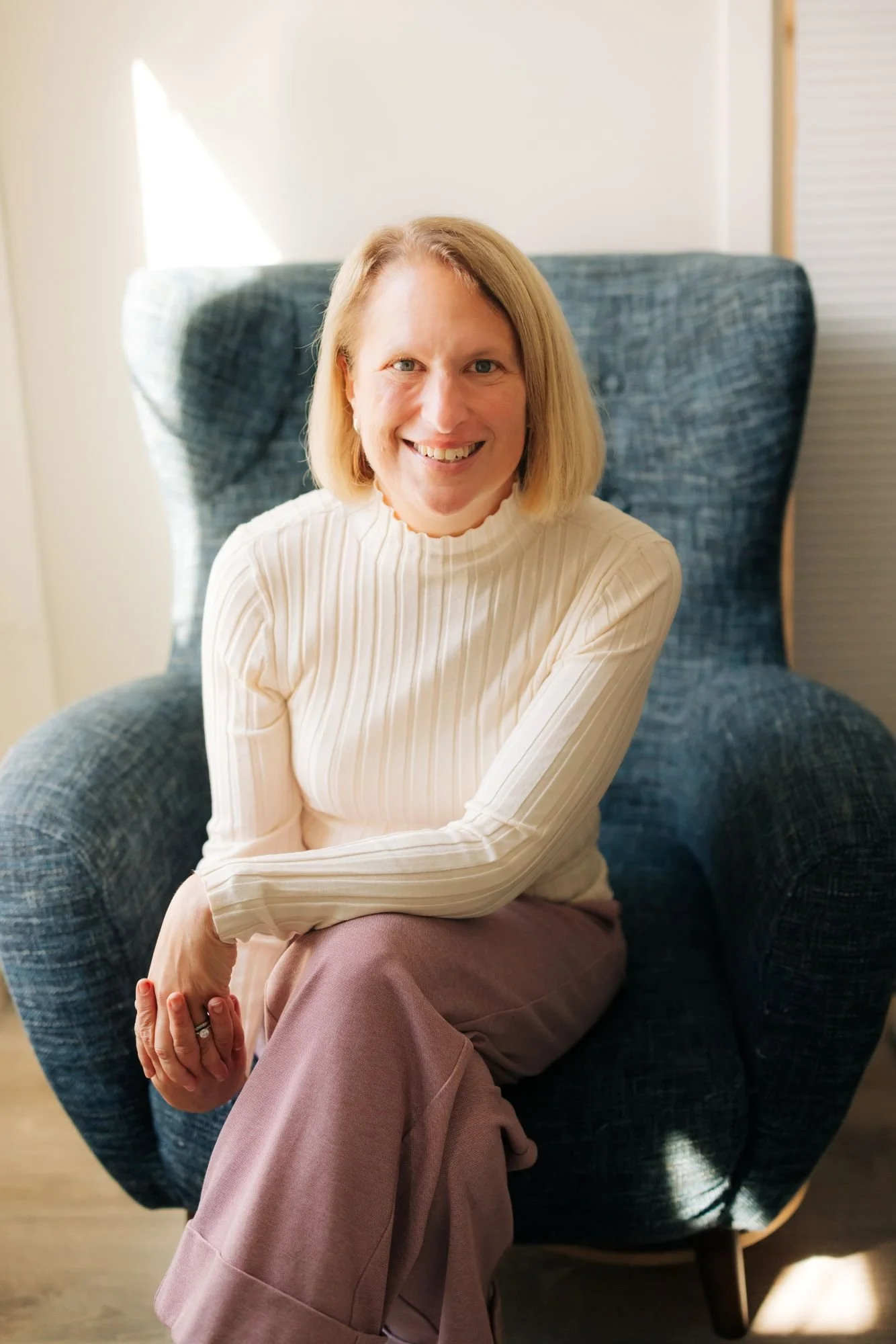 A woman with blonde hair, wearing a cream-colored ribbed turtleneck sweater and pink pants, sitting on a high-back blue armchair in a room with natural light.