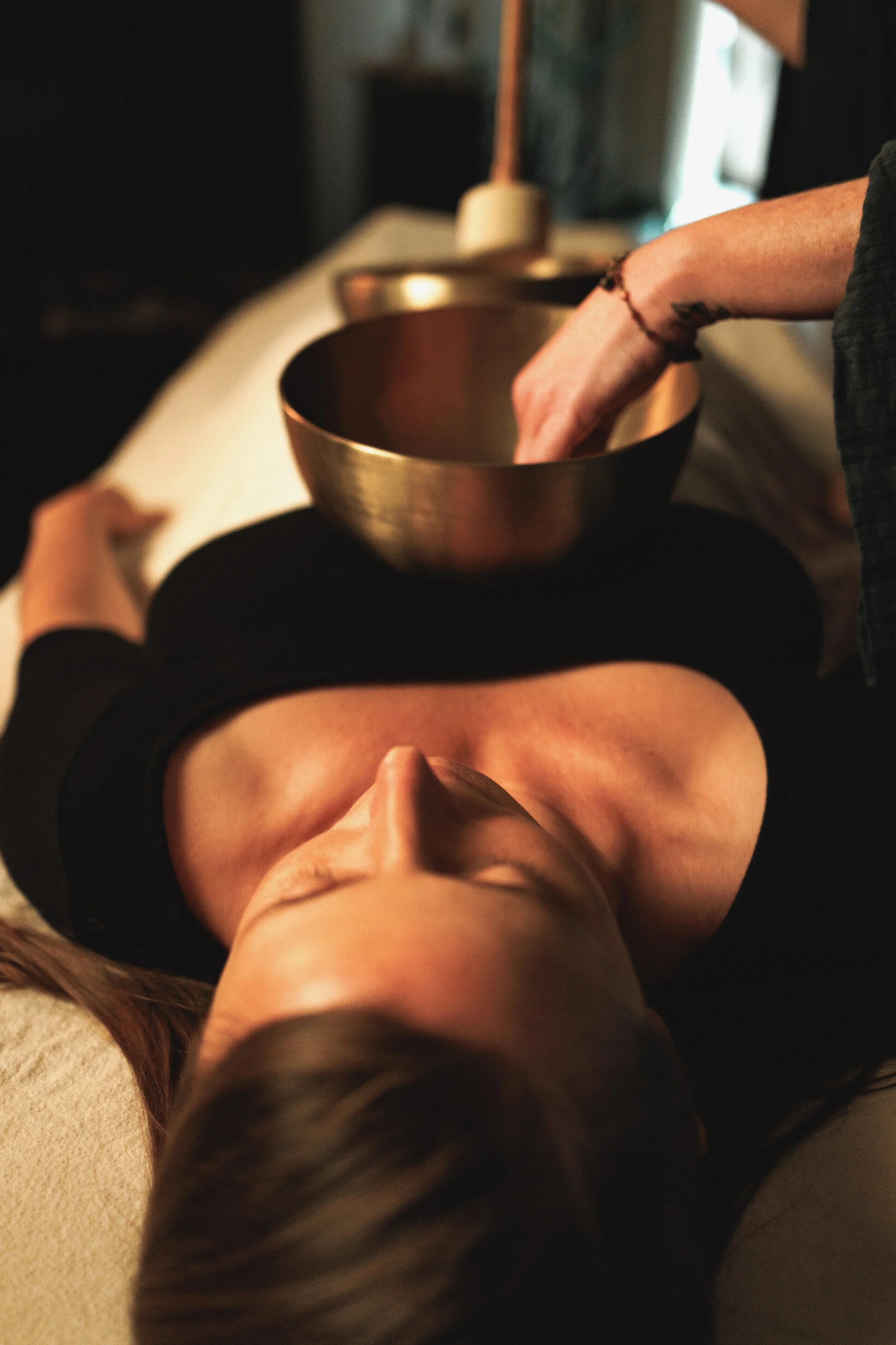 A peaceful Seattle vibrational sound therapy session, featuring a woman resting comfortably on a massage table with singing bowls on her body.  A beautiful and powerful female sound practitioner works with the singing bowls-creating deep relaxation.