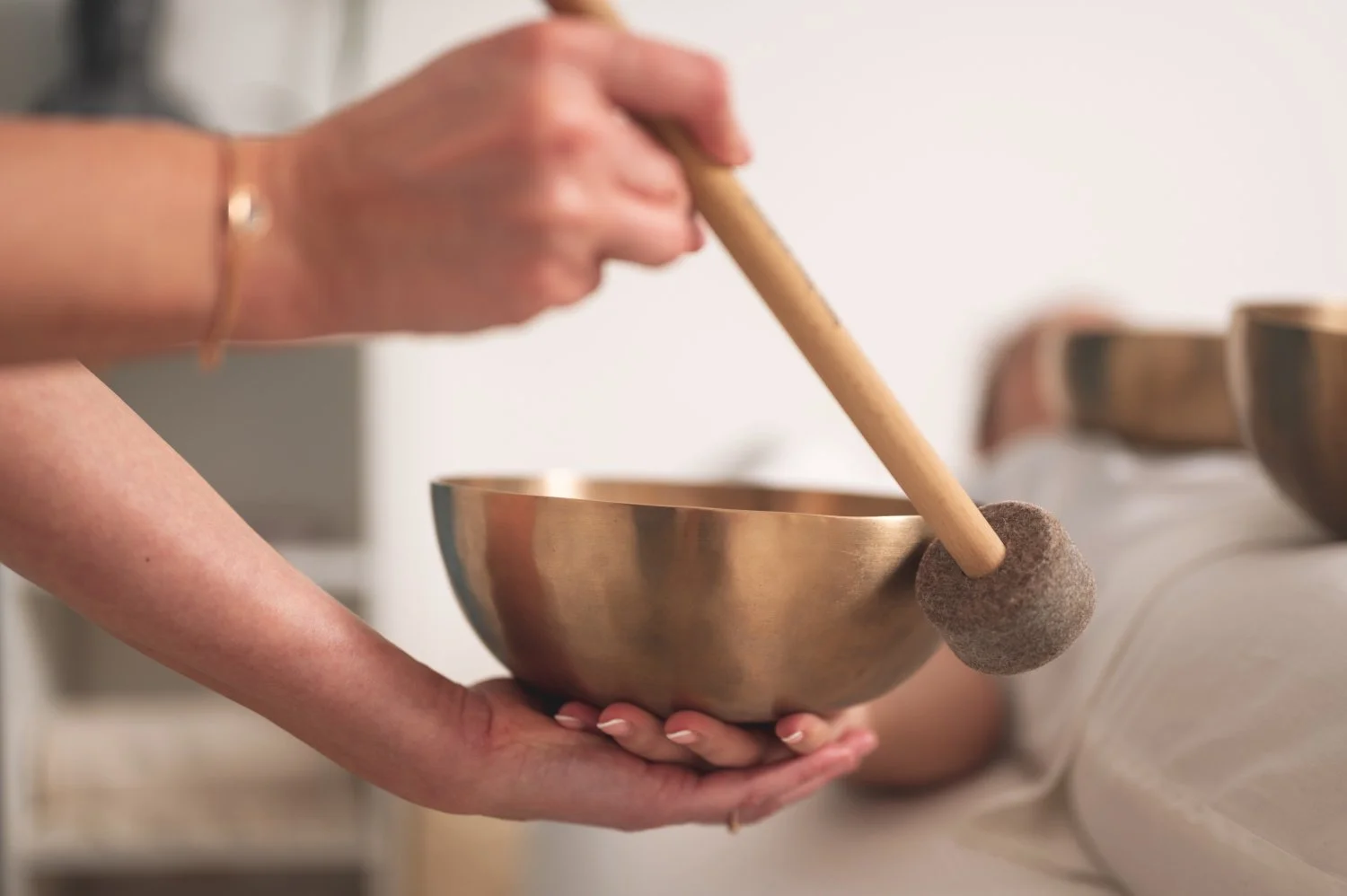 A person holding a singing bowl with one hand while striking it with a mallet in a calm, serene setting.