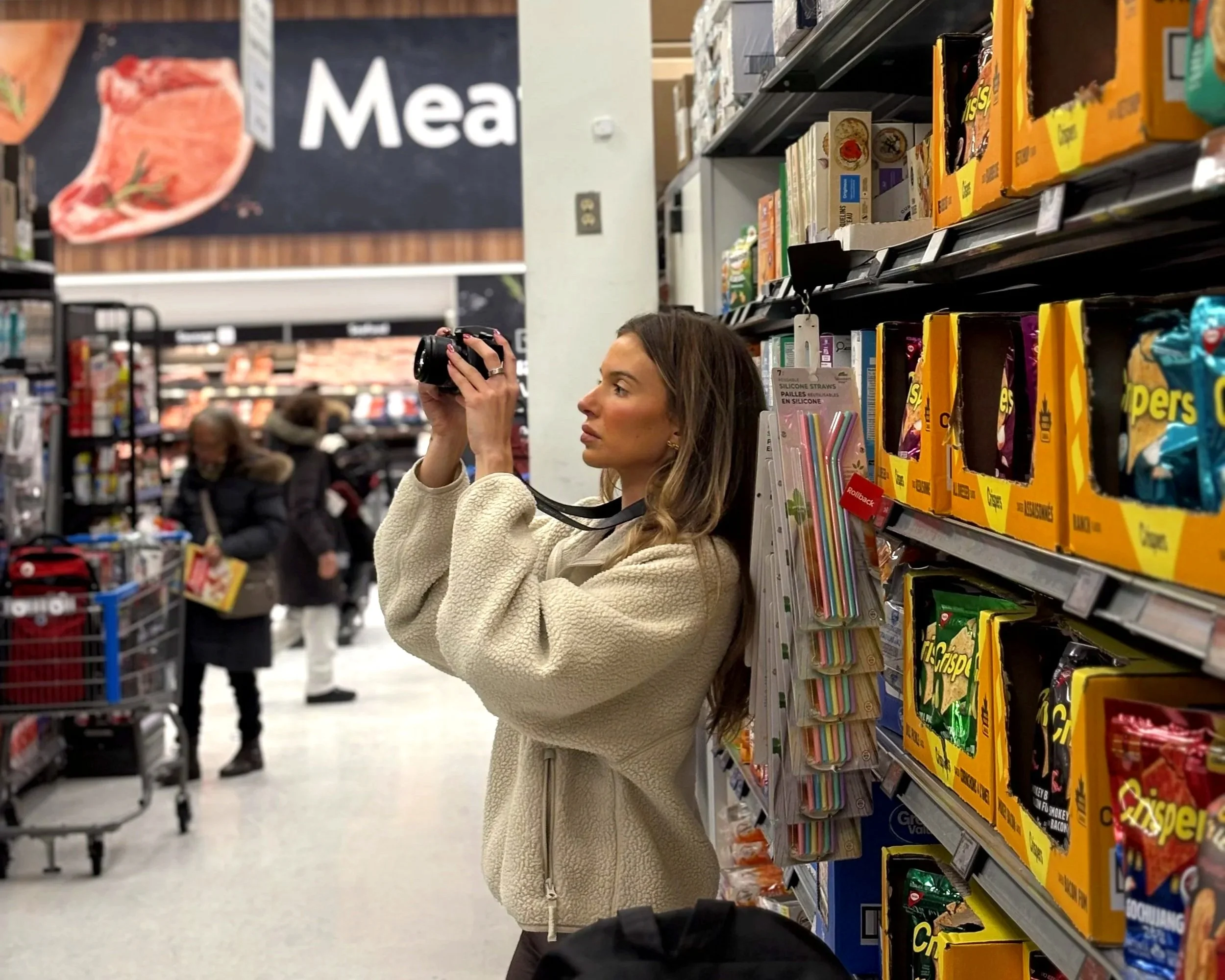 Woman standing in a grocery store aisle taking a picture with a camera.