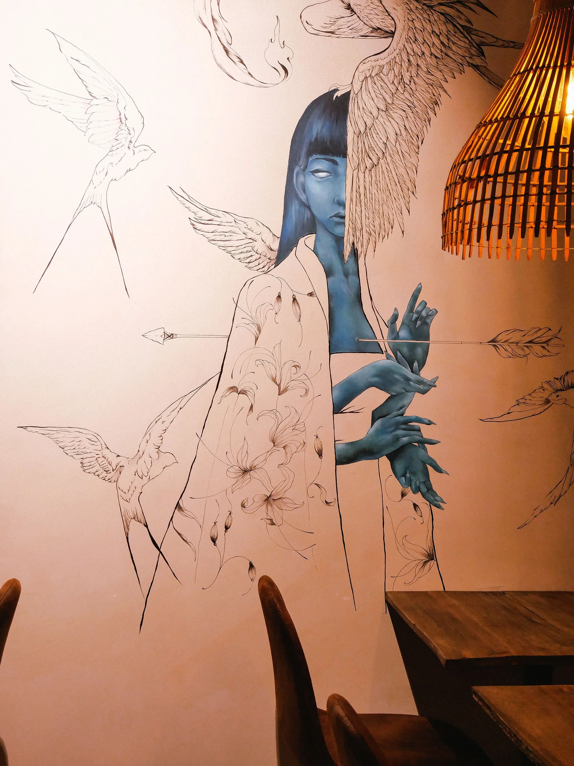 Wall mural of a stylized woman with blue skin, long dark hair, surrounded by flying birds, with some parts in sketch form. A woven hanging lamp is on the right.