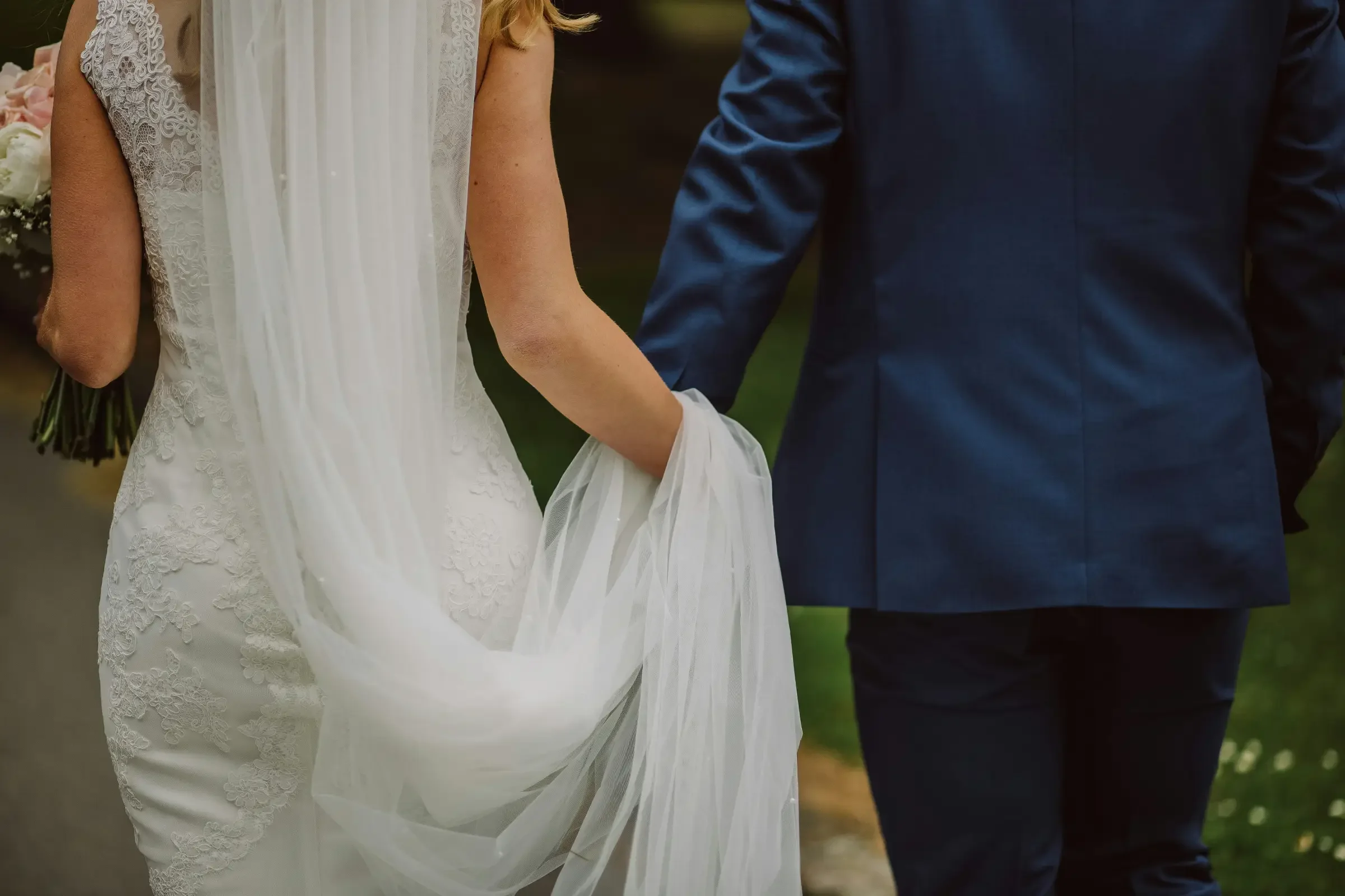 Newlyweds walking through the grounds at Grasshopper Pines, with the bride holding her lace wedding dress train.