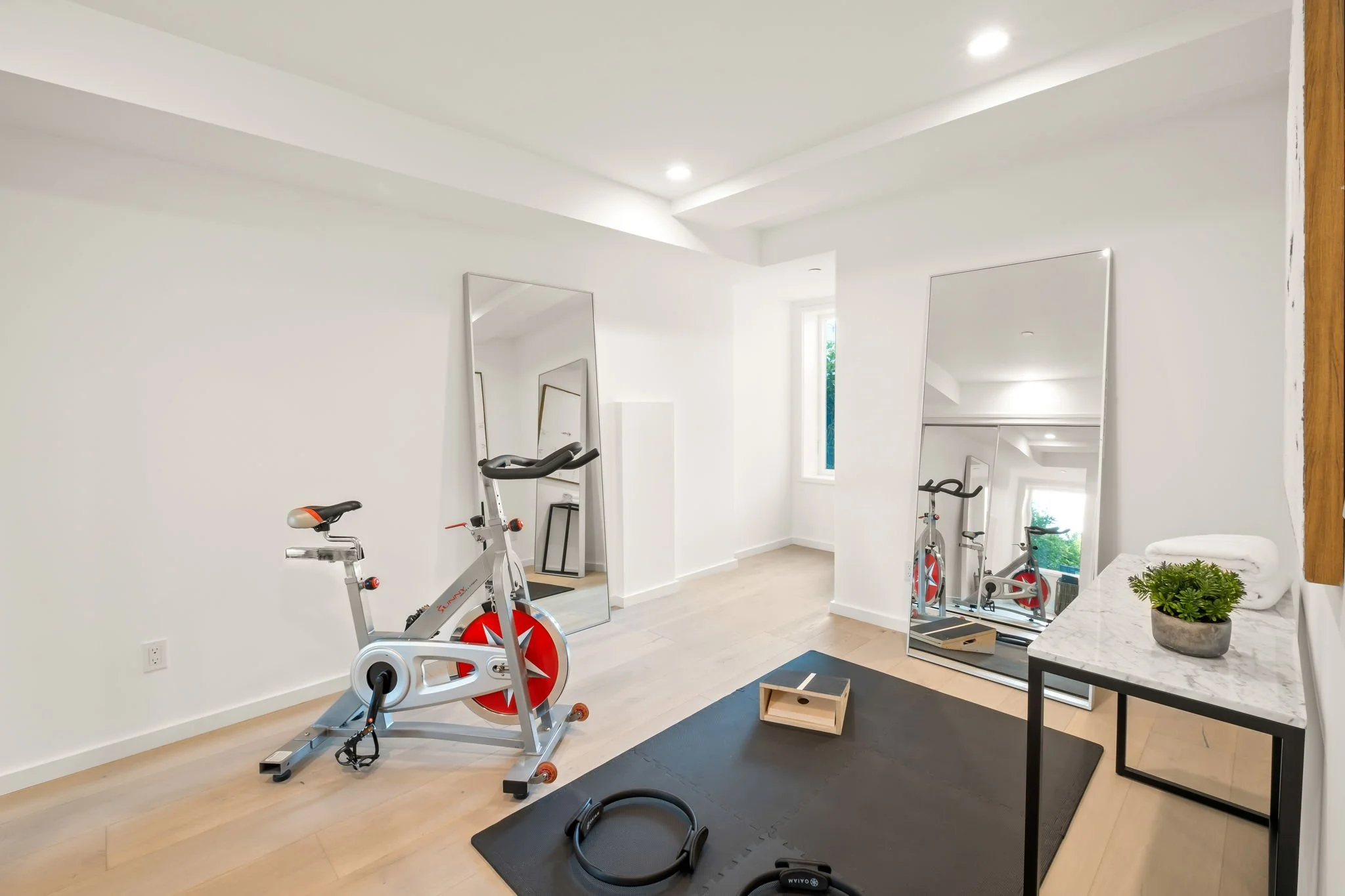 Home gym with a stationary bike, gym mat, mirror, window, black table with potted plant, and fitness equipment.