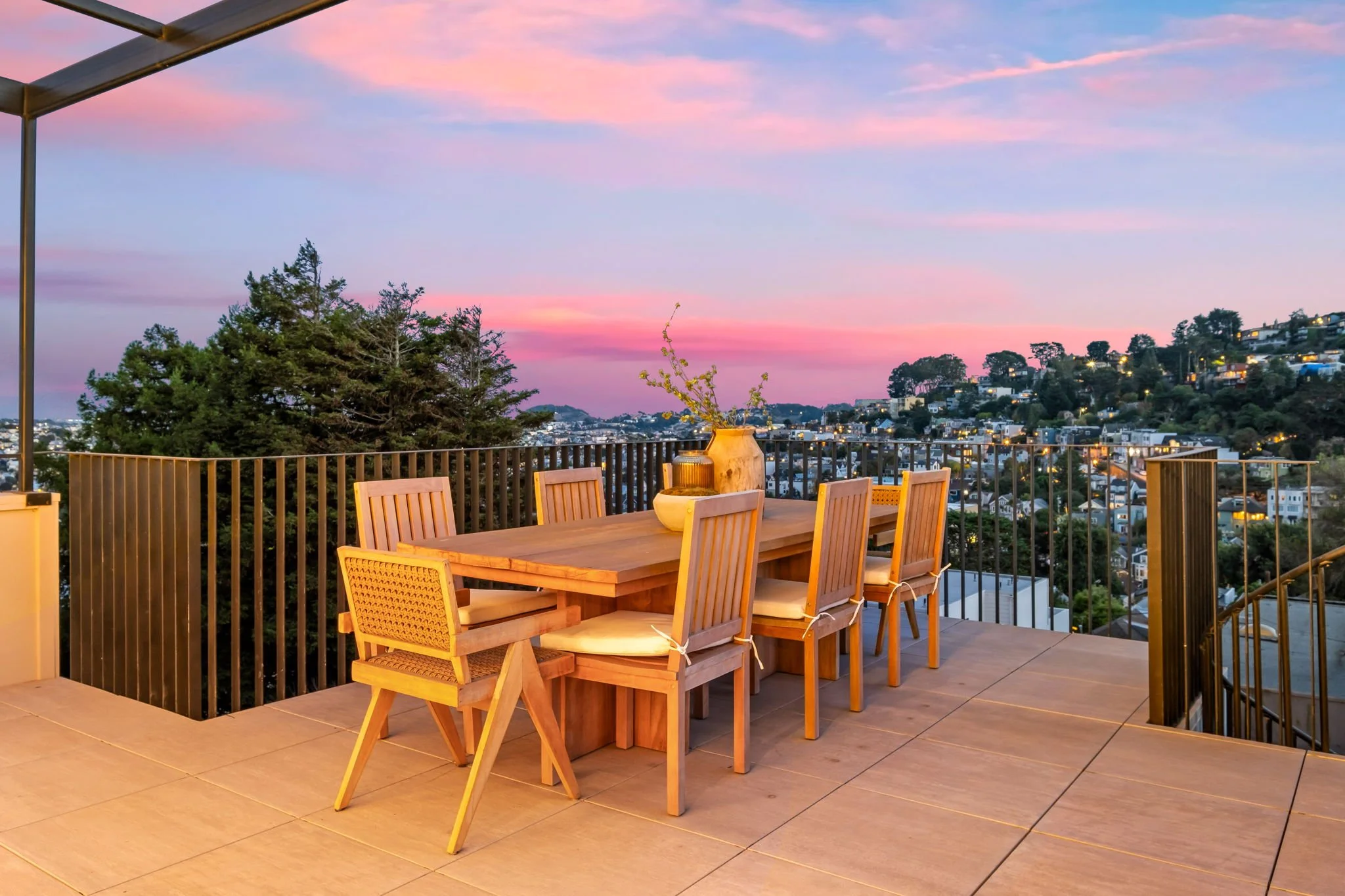 Outdoor dining area on a balcony during sunset with a wooden table, chairs, and a view of a city hillside with houses and trees.