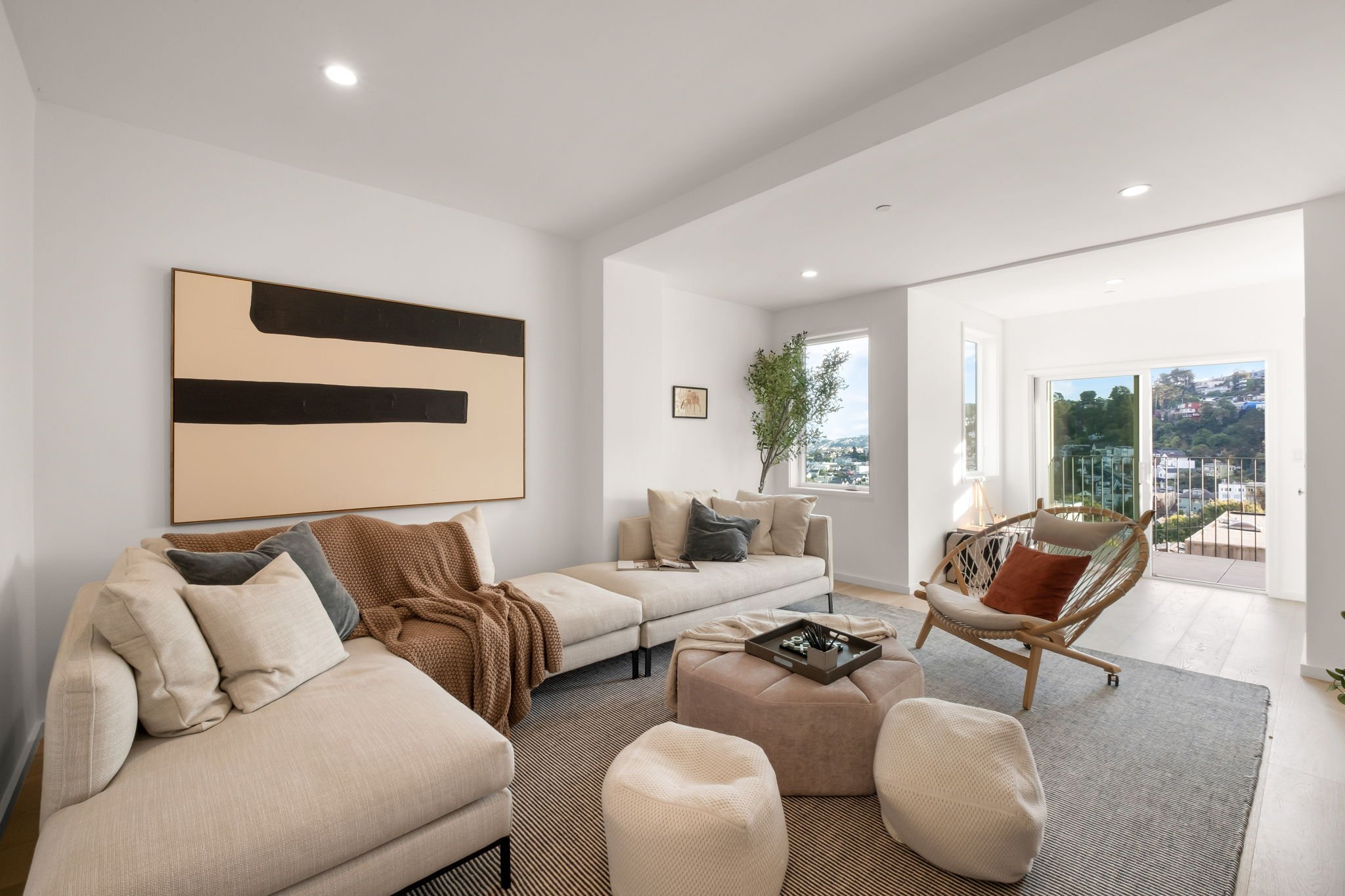Modern living room with white walls, large sofa with beige and gray cushions, abstract artwork, a round ottoman with tray, a woven armchair with a pillow, large windows with a view, and a balcony door.
