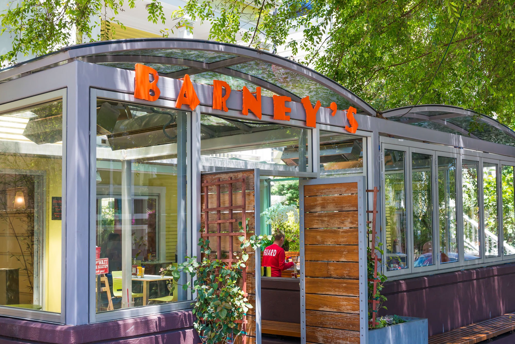 Exterior view of Barney's restaurant with glass walls and a wooden door, surrounded by trees and greenery.