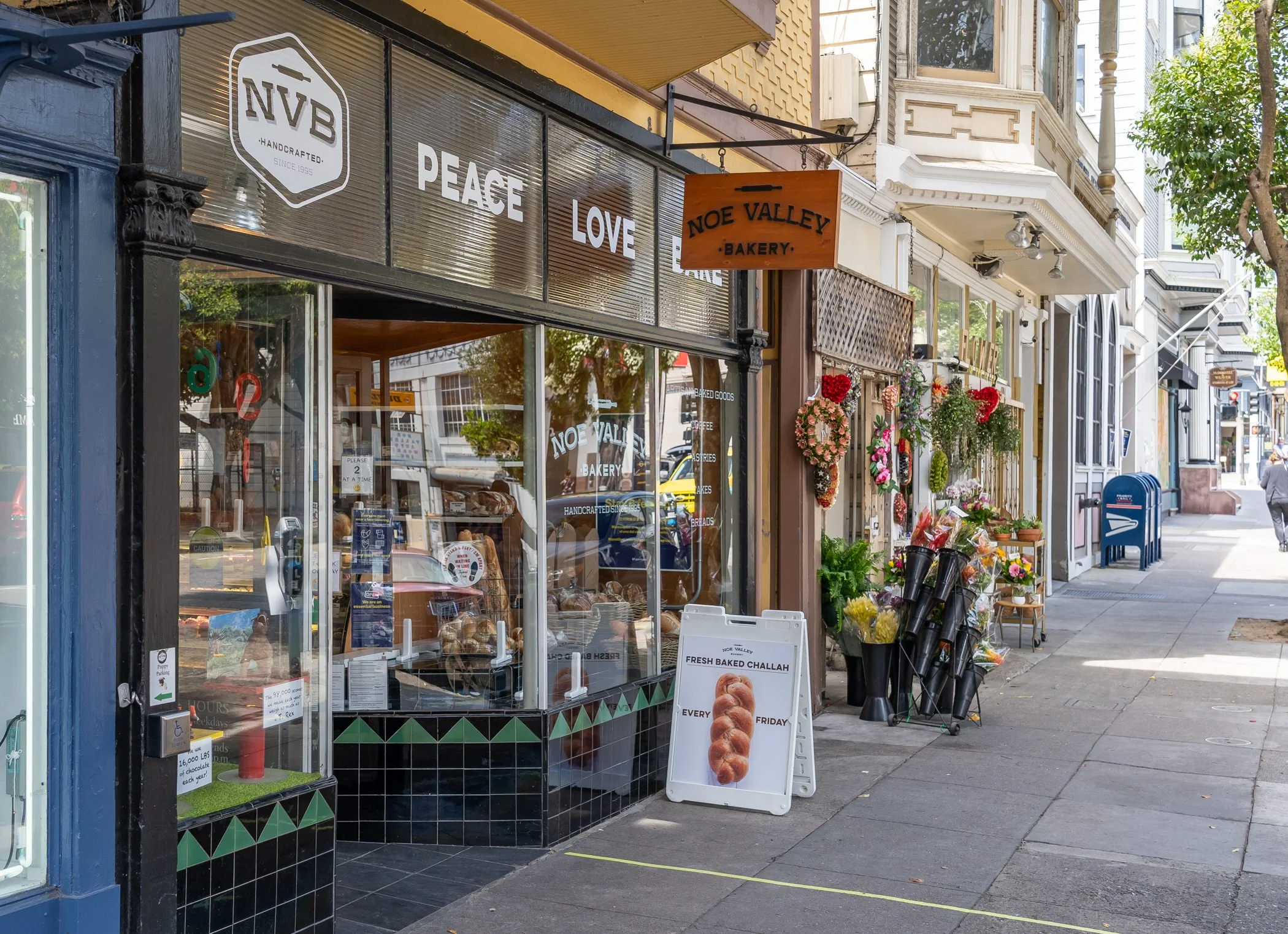 Street view of Noe Valley Bakery storefront with a sign advertising fresh baked challah. There's a display of flowers and plants next to the entrance on a city sidewalk.