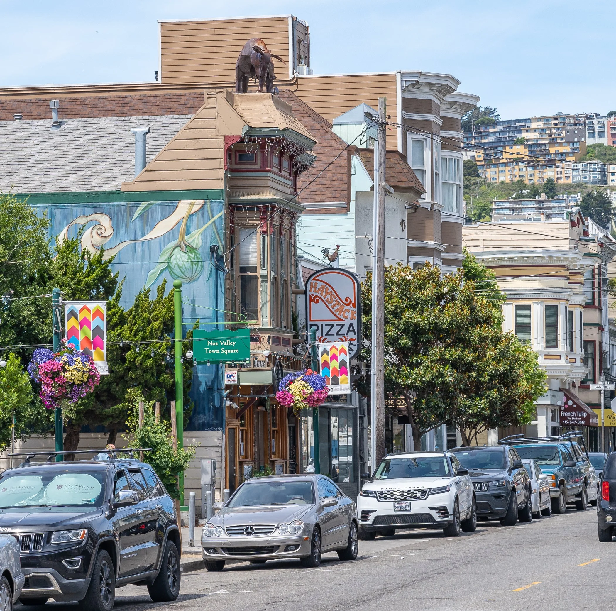 Street view of a neighborhood with colorful buildings, hanging flower baskets, parked cars, and a mural on a building wall featuring a whimsical character, with hillside homes in the background.
