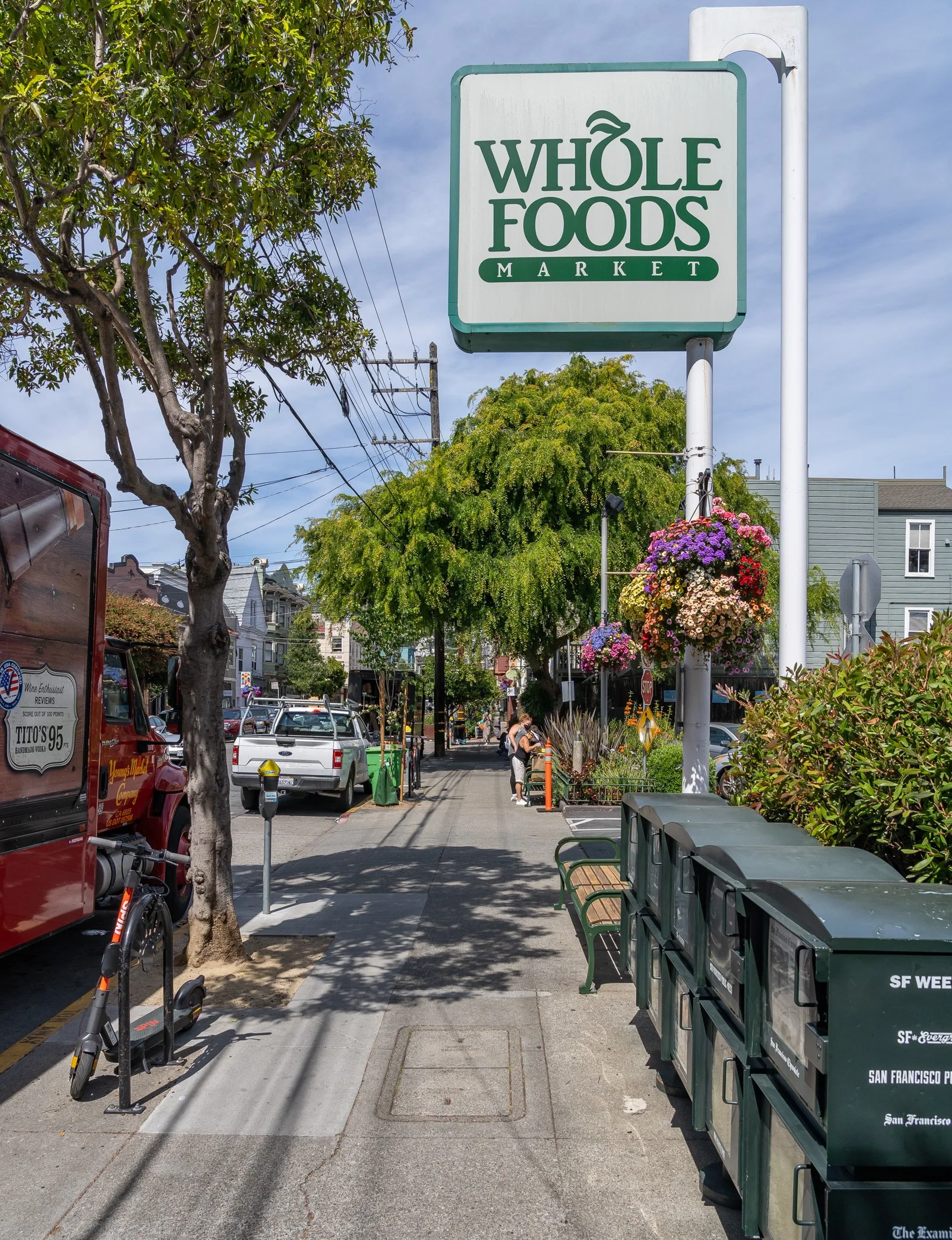 Whole Foods Market sign on a sidewalk with parked vehicles, trees, and newspaper vending boxes.