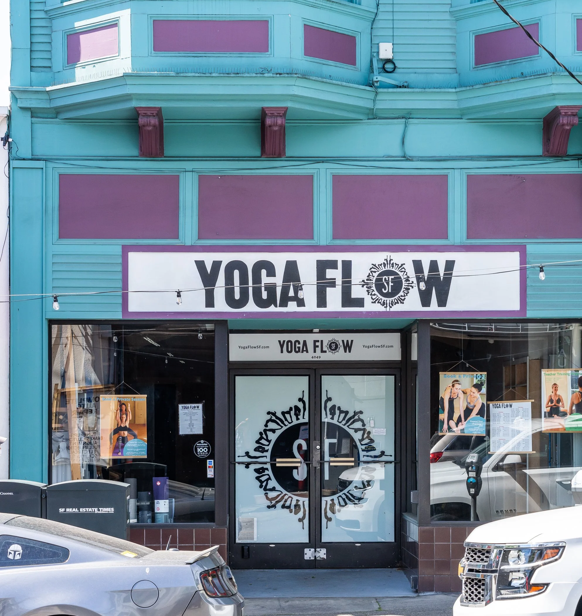 Exterior of a yoga studio with a sign reading 'Yoga Flow SF' above the entrance, featuring turquoise and purple facade. Large windows display yoga-related posters, and cars are parked in front of the building.
