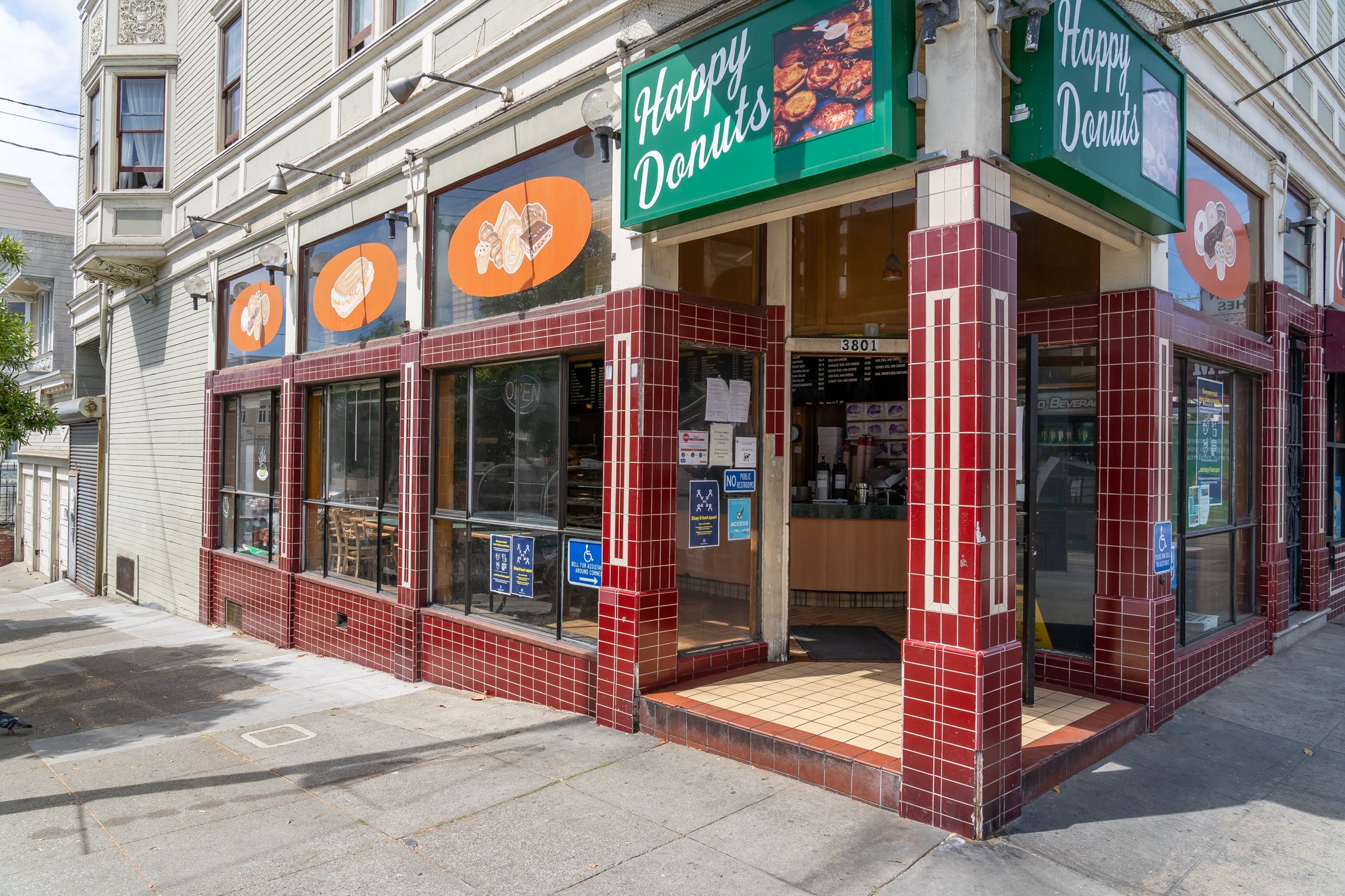 Exterior of "Happy Donuts" shop on a corner, with red tiled facade and large windows. Bright green sign above entrance. Sidewalk and street visible outside, signs indicating accessibility.