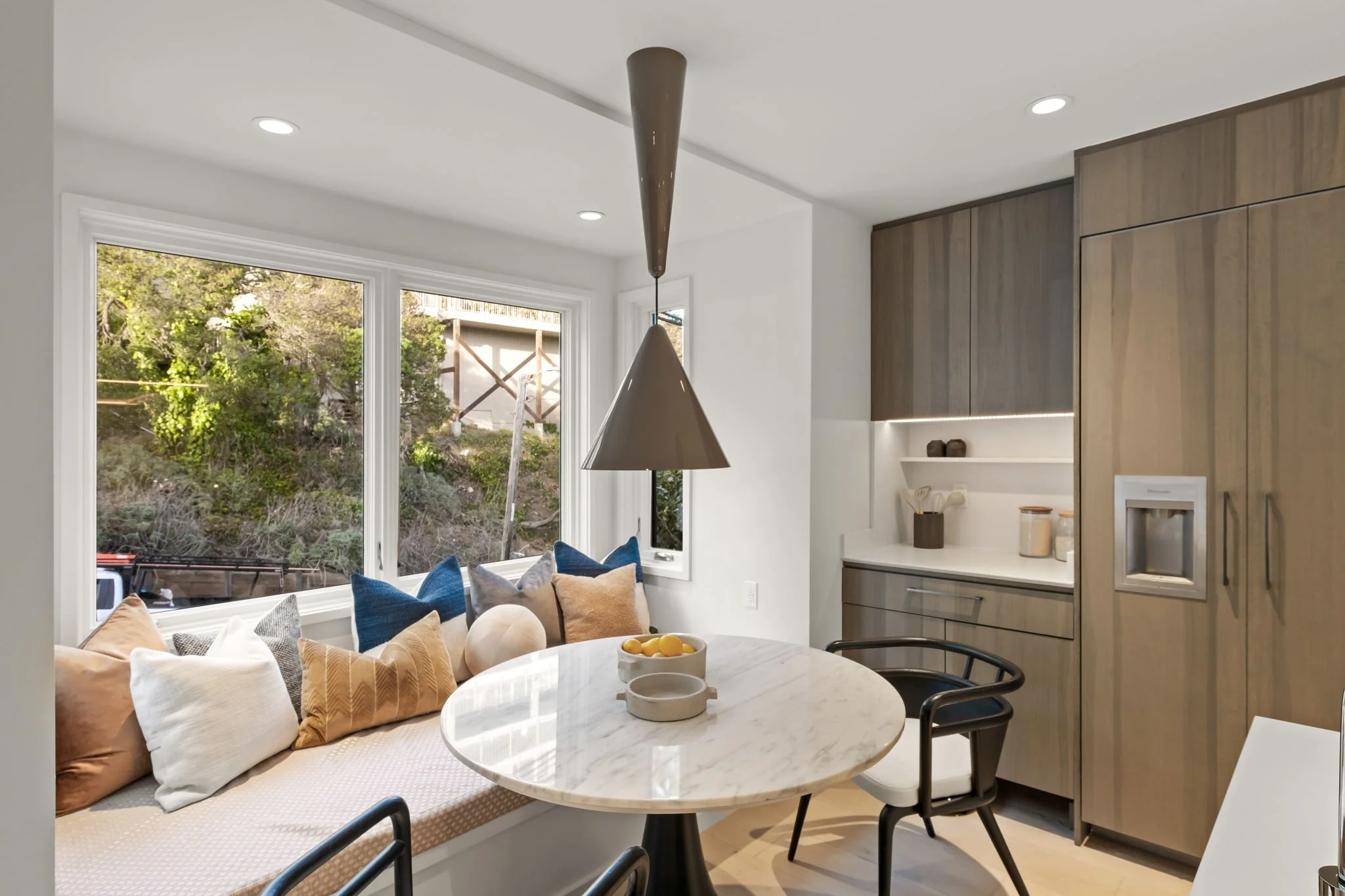 Modern kitchen with large window seat decorated with pillows, a marble dining table, and wooden cabinetry with built-in appliances.