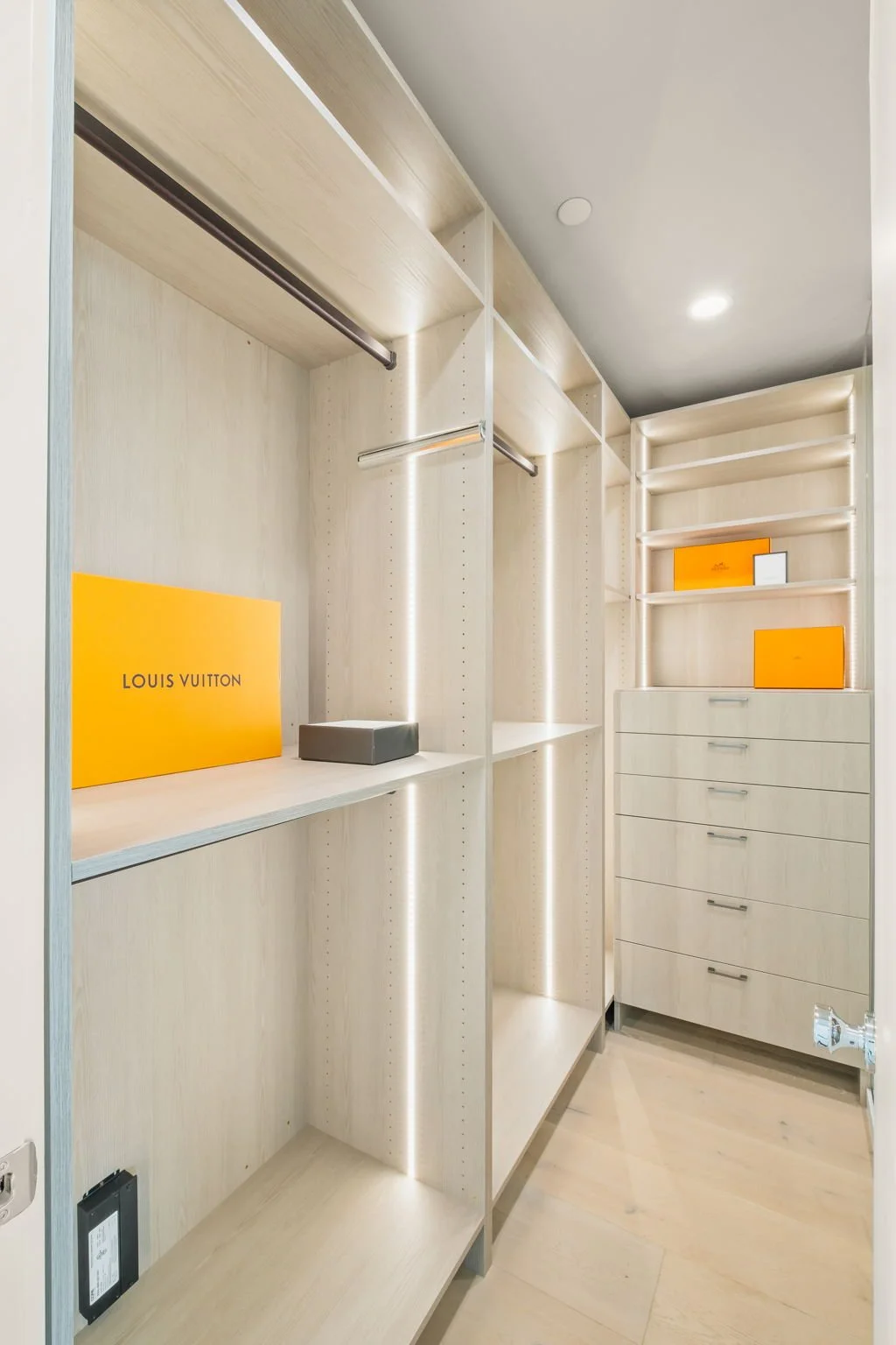 Modern walk-in closet with light wood shelves, drawers, and jewelry display. Contains orange Louis Vuitton boxes and a black accessory box.