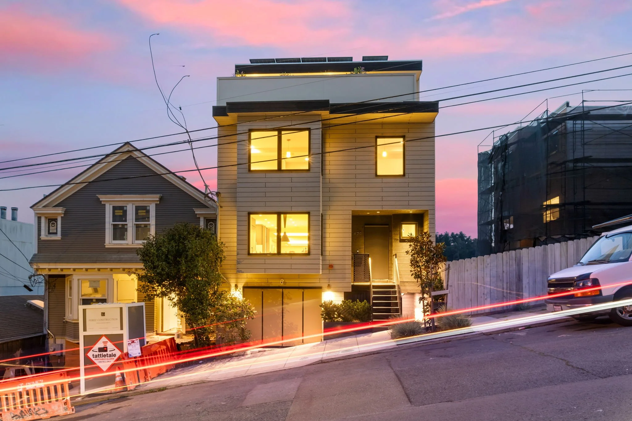 A modern three-story house with illuminated windows, situated on a sloped street during dusk. The sky is pink and purple, with neighboring houses and construction scaffolding visible.