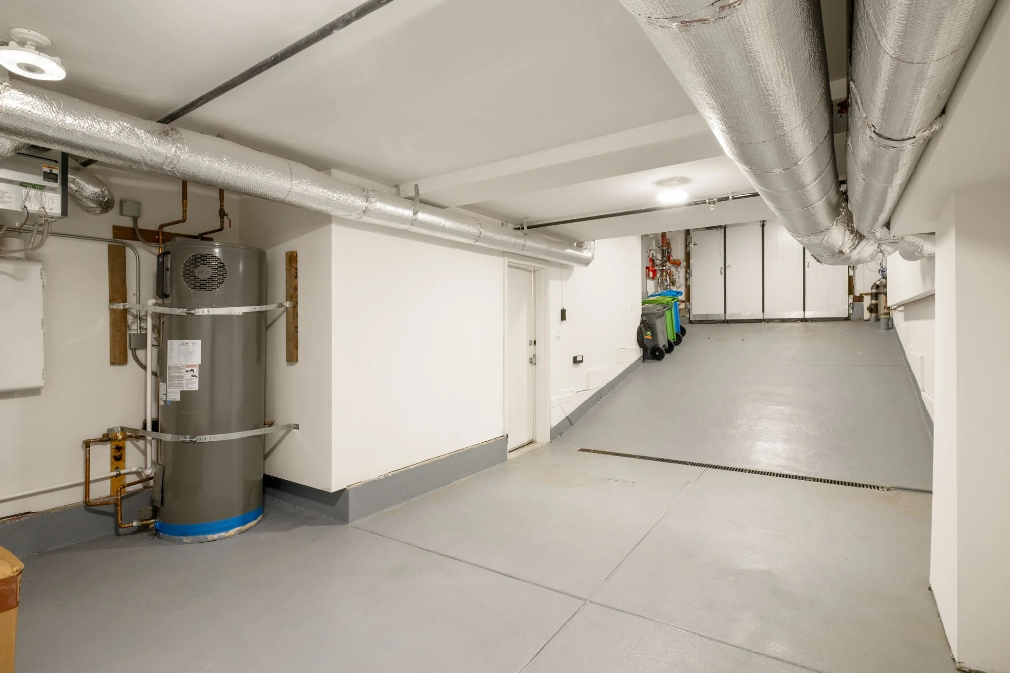 Empty garage with a water heater, pipes, and a few trash bins against the wall.