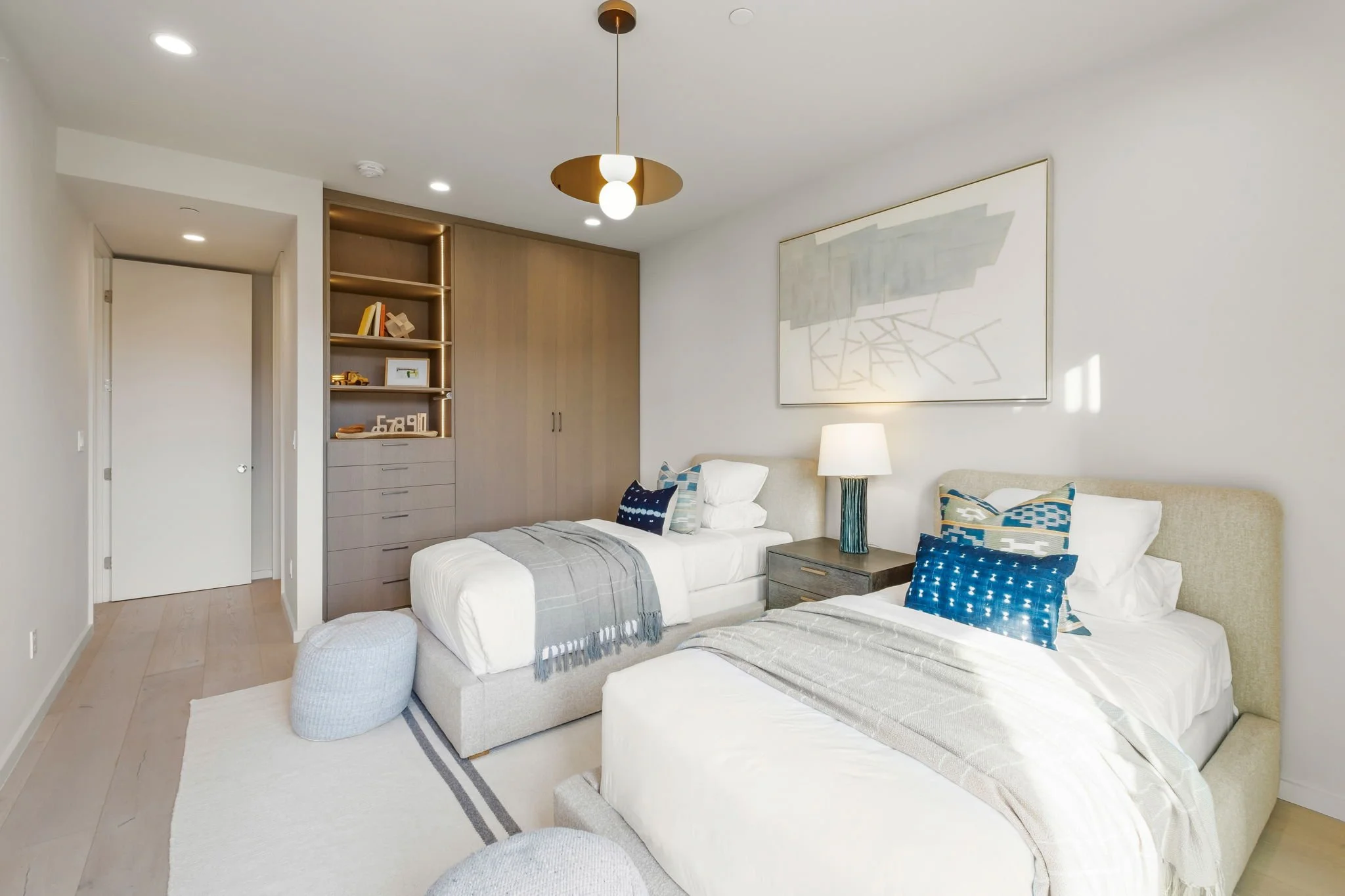 Modern bedroom with two beds, decorative pillows, white and gray bedding, wooden nightstand with a lamp, abstract wall art, built-in wooden closet, and light-colored hardwood flooring.