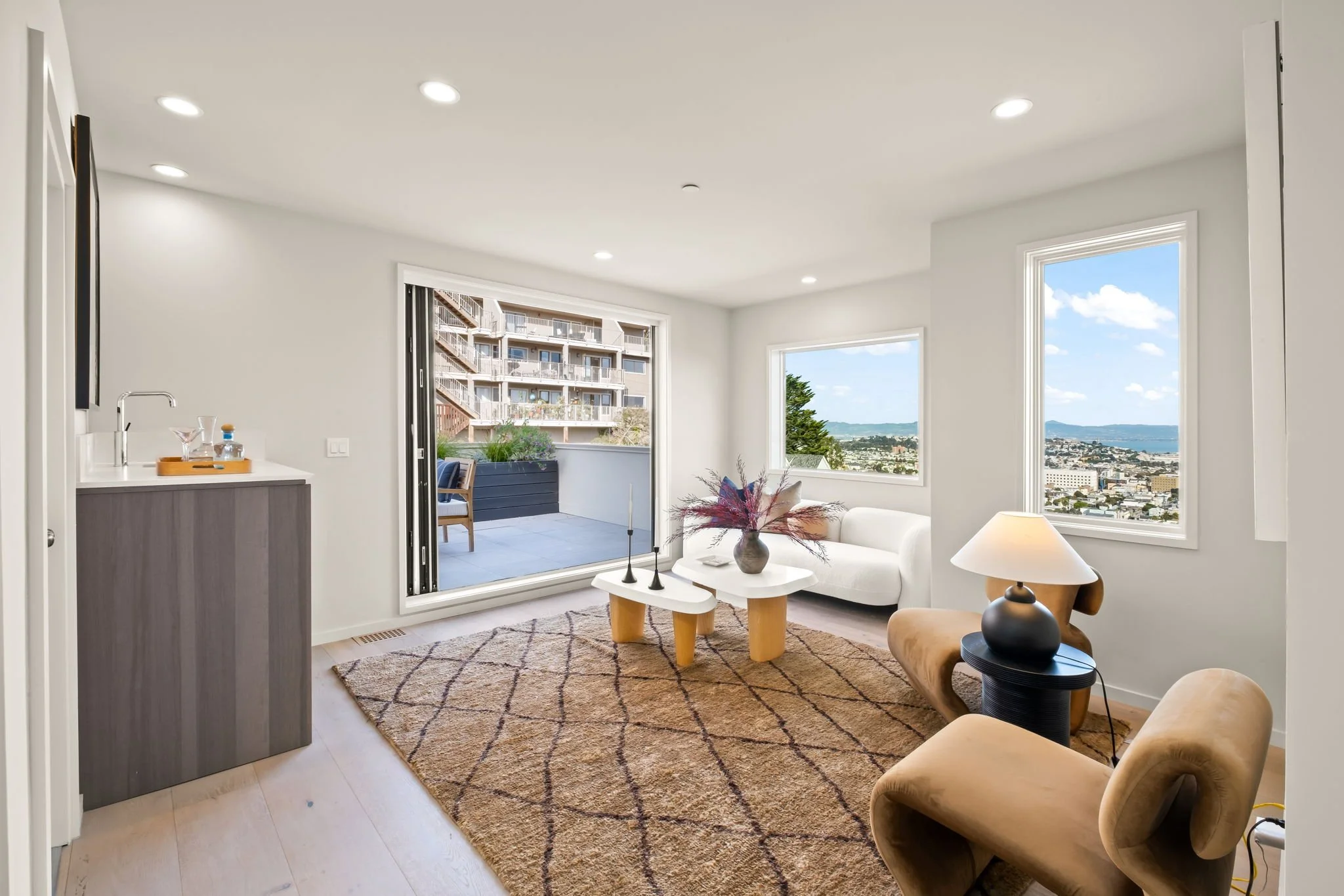 Living room with a white sofa, beige armchair, coffee table with decorative items, large windows showing city and ocean views, sliding door to balcony with outdoor furniture, area rug, and a side table with a lamp.