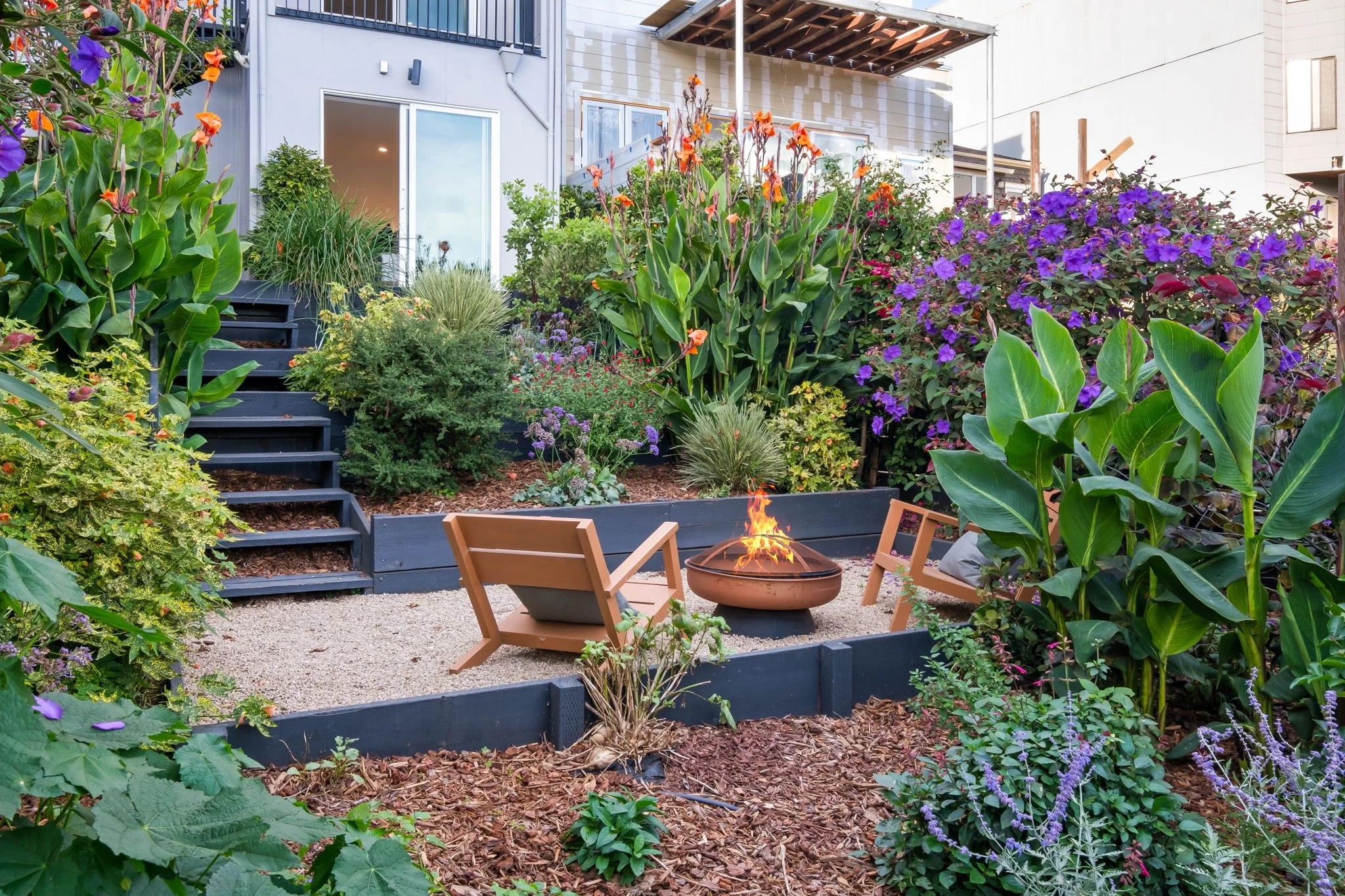 Urban backyard garden with colorful flowering plants, a fire pit with flames, and wooden chairs, overlooking a modern house with a glass door.