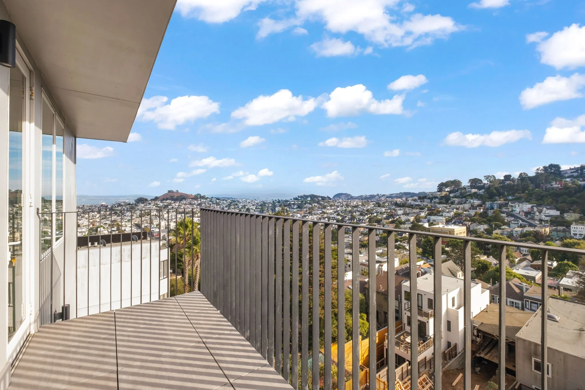 View from a balcony overlooking a cityscape with hills, trees, and scattered houses under a blue sky with clouds.