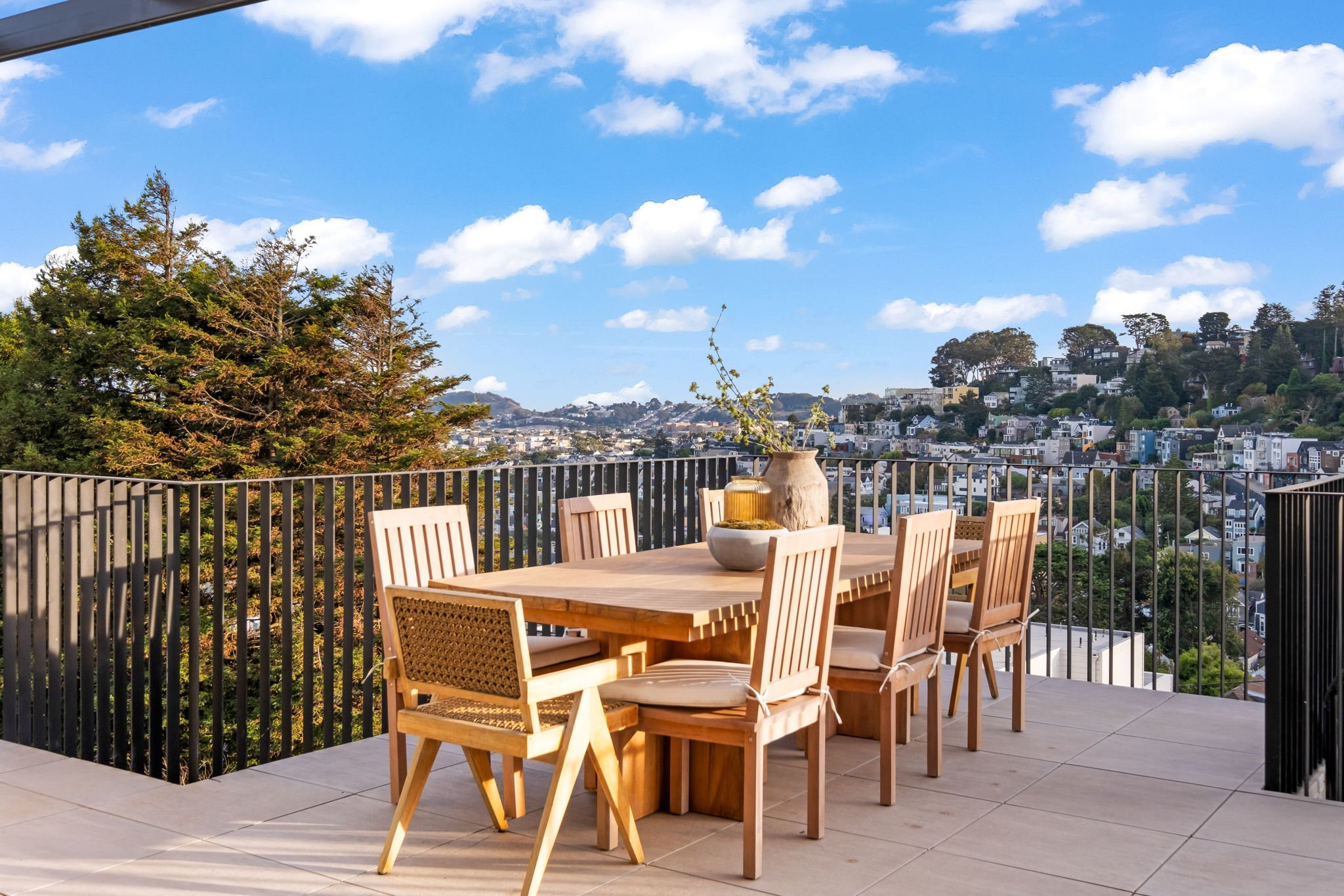 Outdoor patio with a wooden dining table and six chairs, decorated with vases and plants, overlooking a cityscape with houses and trees under a blue sky with scattered clouds.