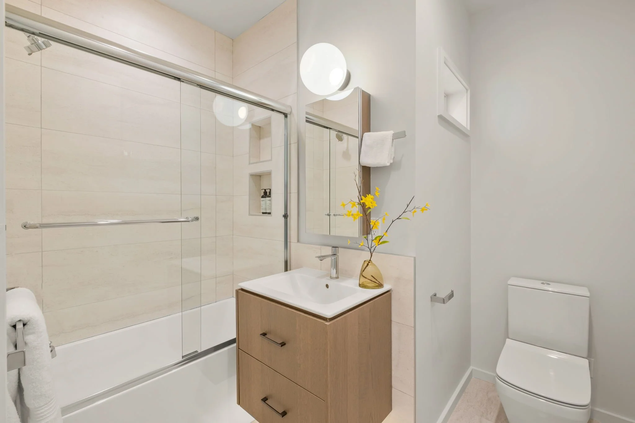 Modern bathroom with beige tiled shower, white toilet, and wooden vanity with sink, mirror, and yellow flowers in a glass vase.
