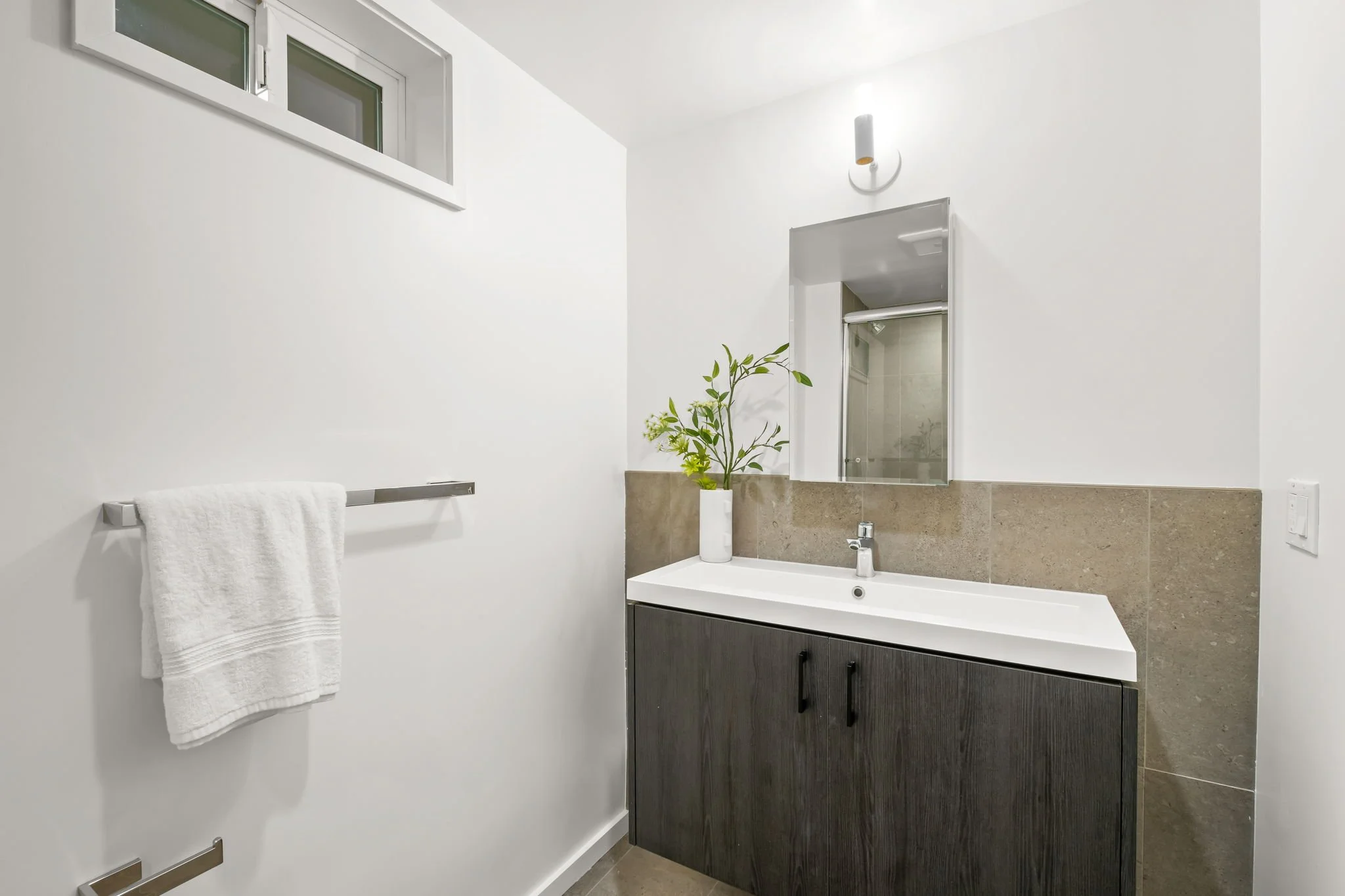 Modern bathroom with white walls, small window, wall-mounted towel rack with white towel, and a sink cabinet with a mirror and a small plant.
