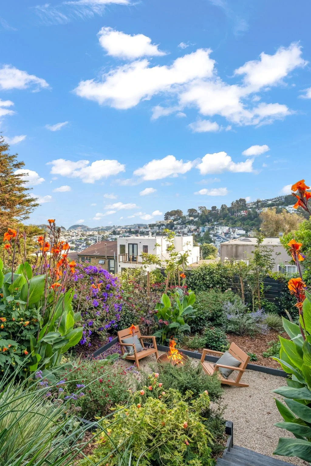 Sunlit garden with flowering plants and two wooden chairs facing a small fire pit, overlooking a hillside neighborhood with houses and trees under a blue sky with scattered clouds.