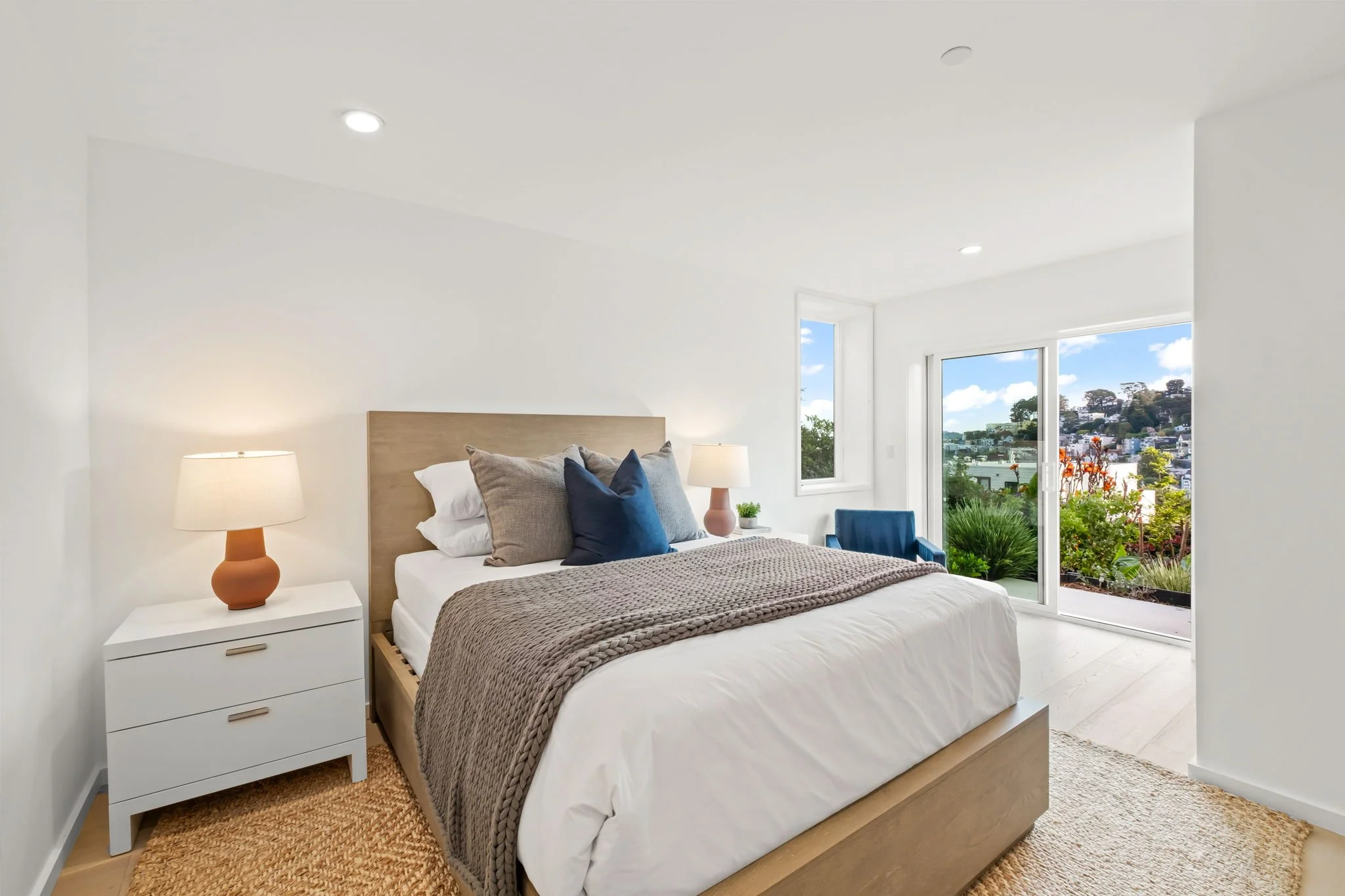 Modern bedroom with white walls, wooden bed frame, beige and navy pillows, knitted throw blanket, white nightstand with terracotta lamp, and large sliding glass door opening to a balcony with greenery and city view.