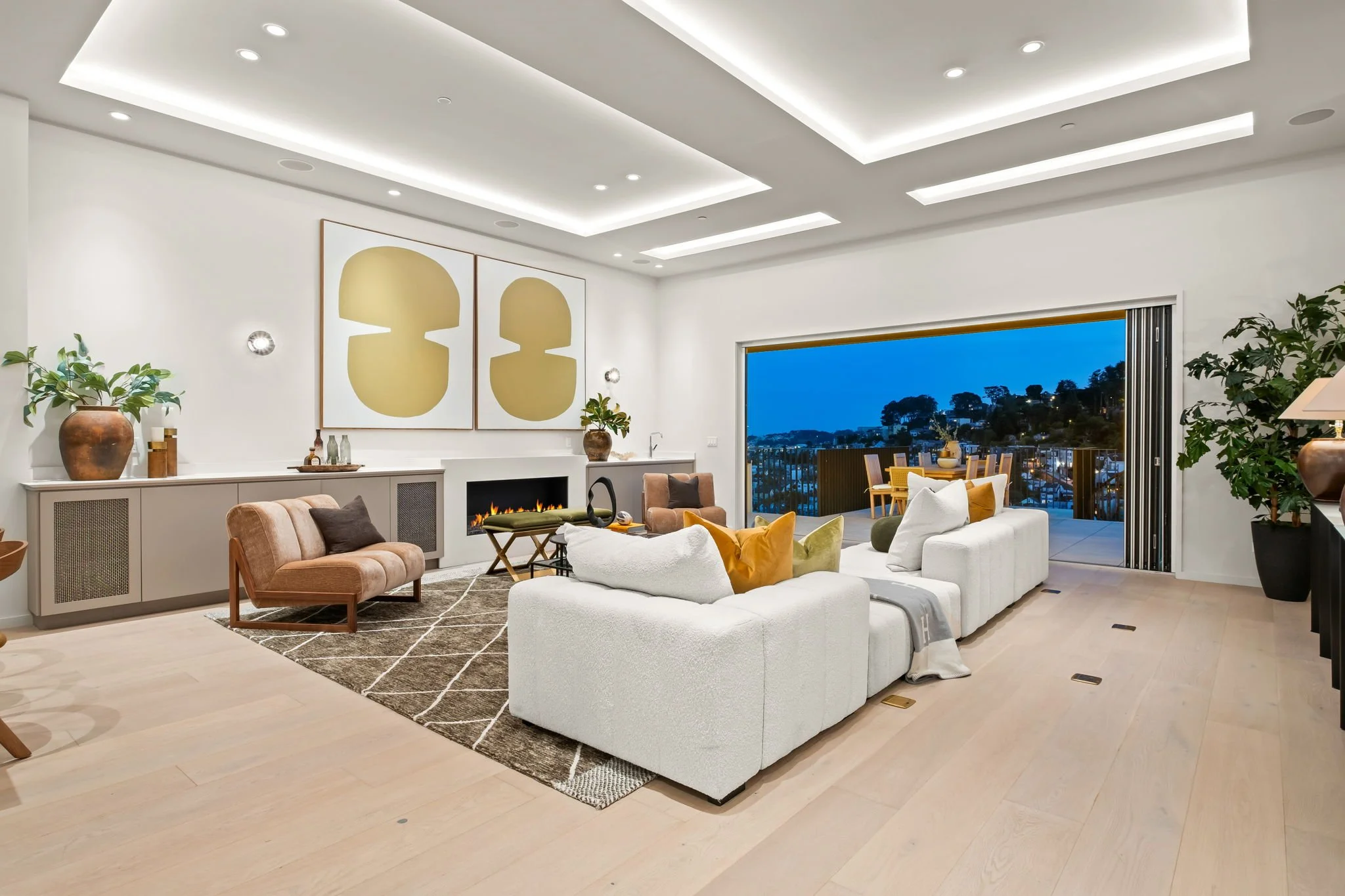 Living room with white sofa, pink armchair, black fireplace, large abstract art, plants, and a sliding door opening to a balcony with outdoor dining, overlooking a hillside at dusk.