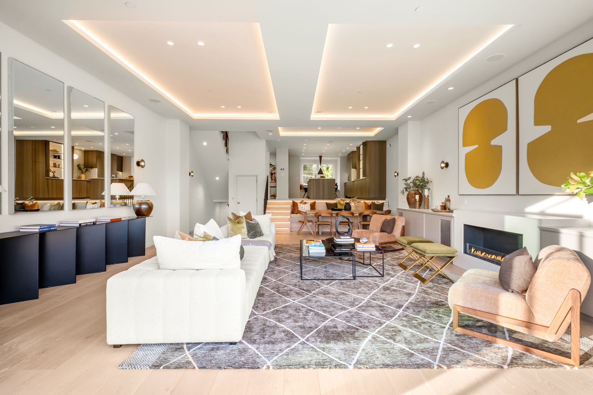 Spacious modern living room with white walls, beige and pastel furniture, a patterned gray and white rug, and a sleek fireplace on the right wall. Decor includes abstract artwork and contemporary accents.