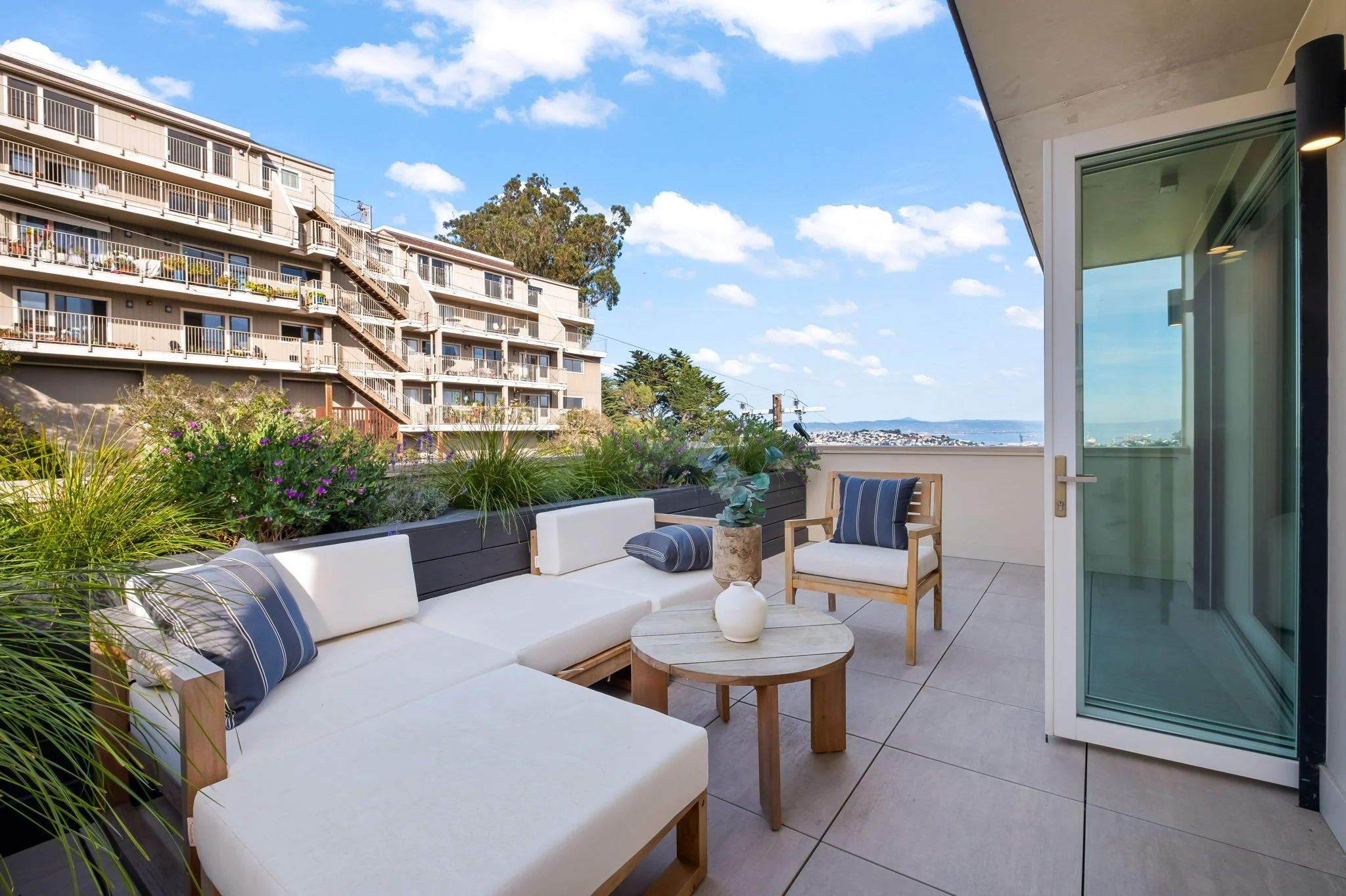 Balcony with white cushioned seating, a wooden armchair with navy striped pillows, a small round wooden table with a white vase and green plant, overlooking neighboring apartment buildings and a view of a city and water under a blue sky with clouds.