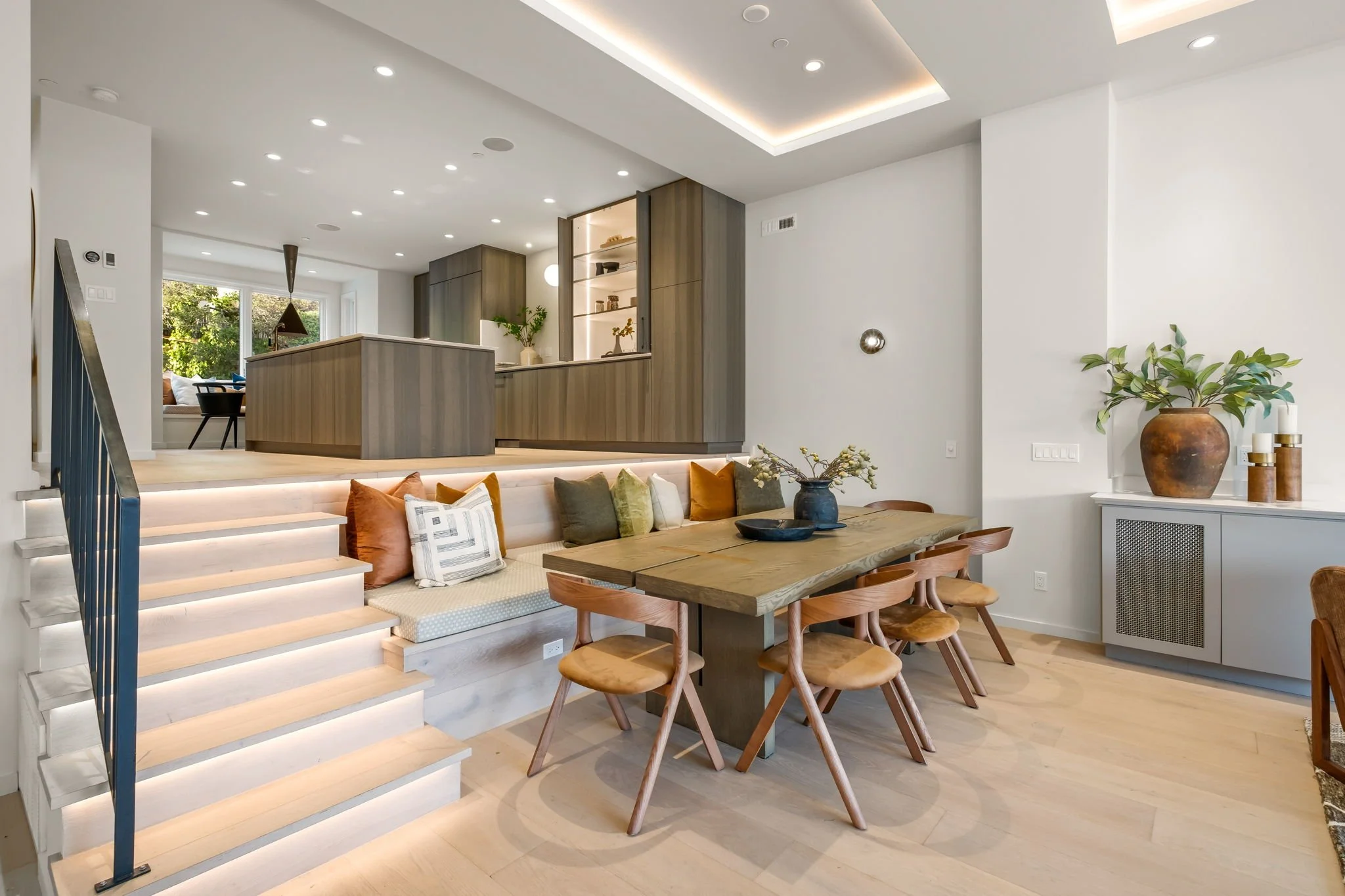 Modern open-concept living space with a dining area, built-in seating with colorful pillows, wooden dining table, and staircase with LED lighting. Kitchen with wooden cabinets and large window in background, decorative plants, and minimalist decor.