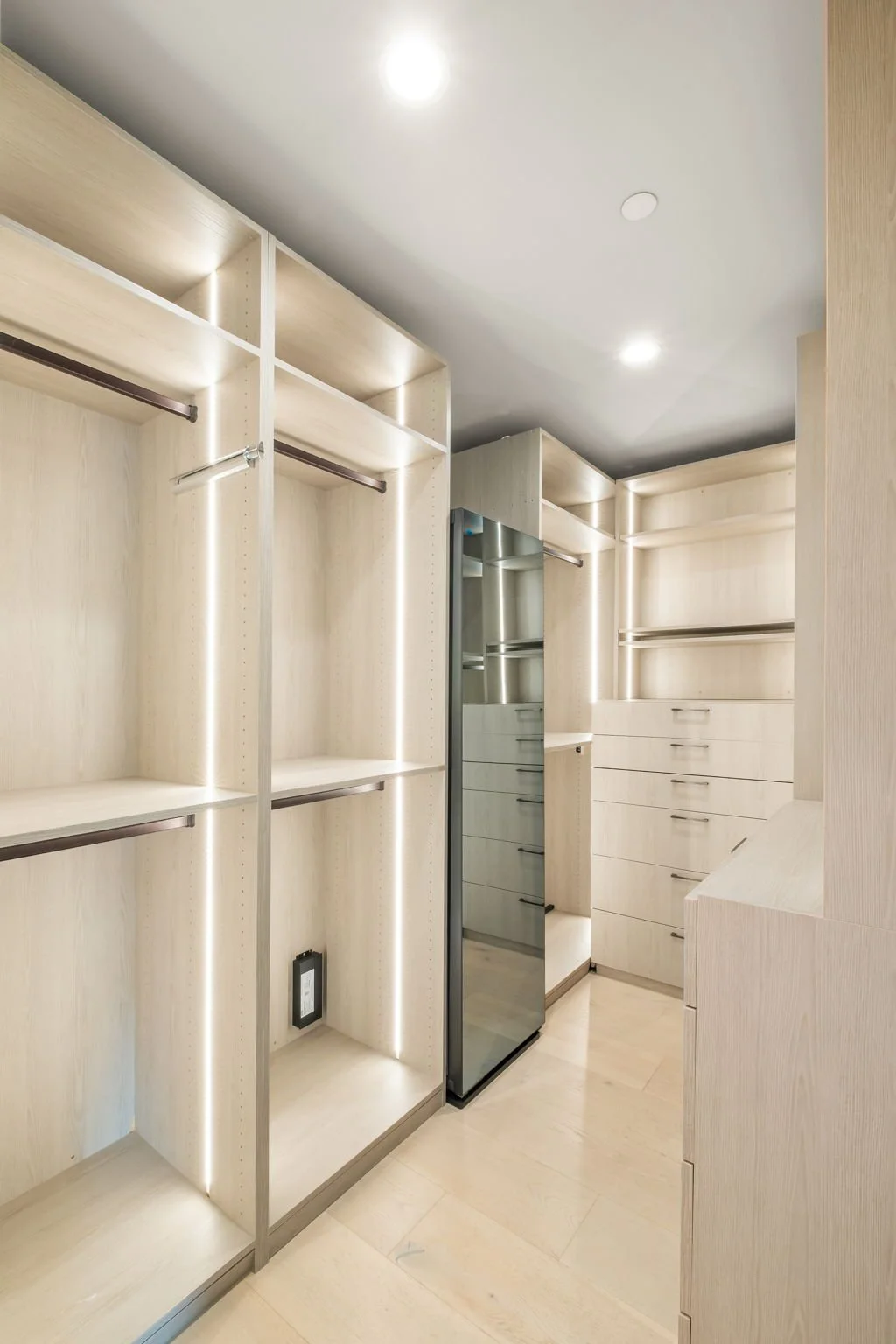 A modern walk-in closet featuring light wood shelving, drawers, and hanging rods, with built-in LED lighting and a mirrored door.