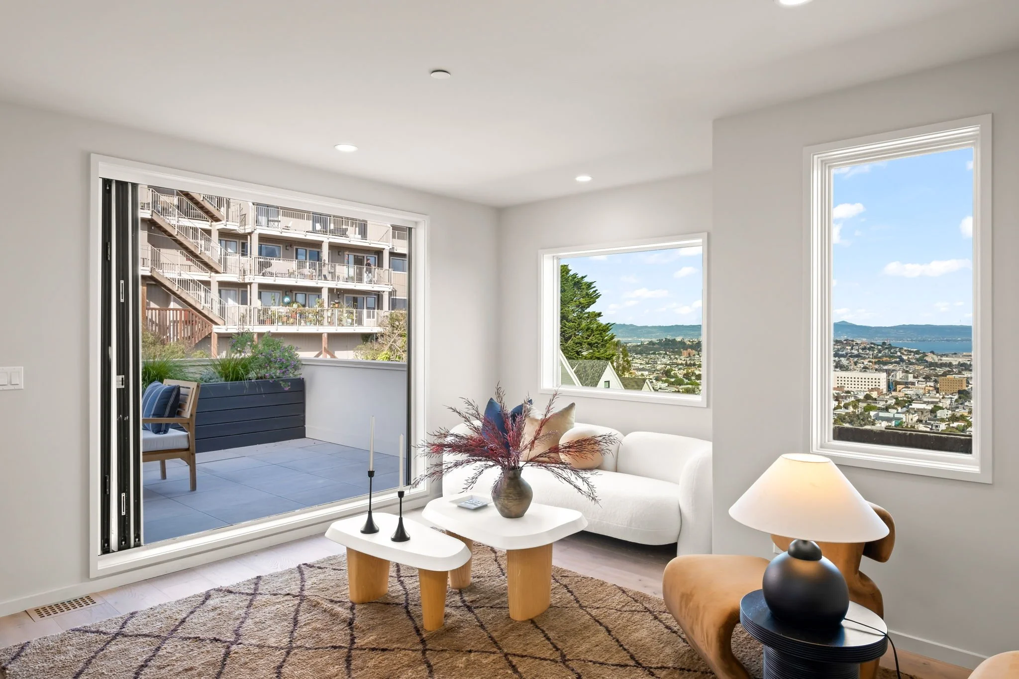 Living room with large windows showing cityscape and water view, white sofa, side table with lamp, and balcony outside.