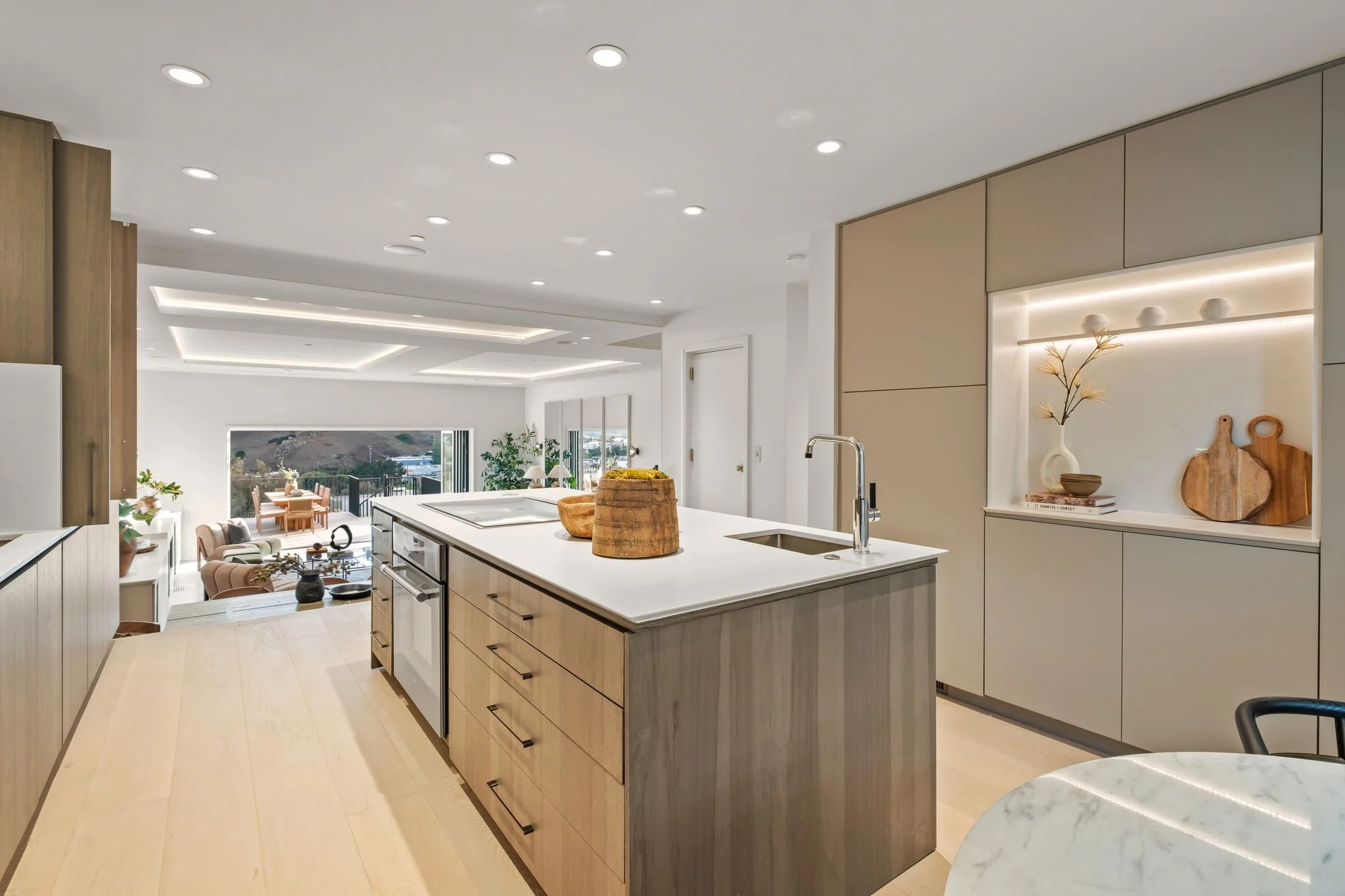Modern kitchen with a white countertop, wooden cabinets, and open shelving showcasing decorative items; view into the living area with large windows and outdoor scenery.