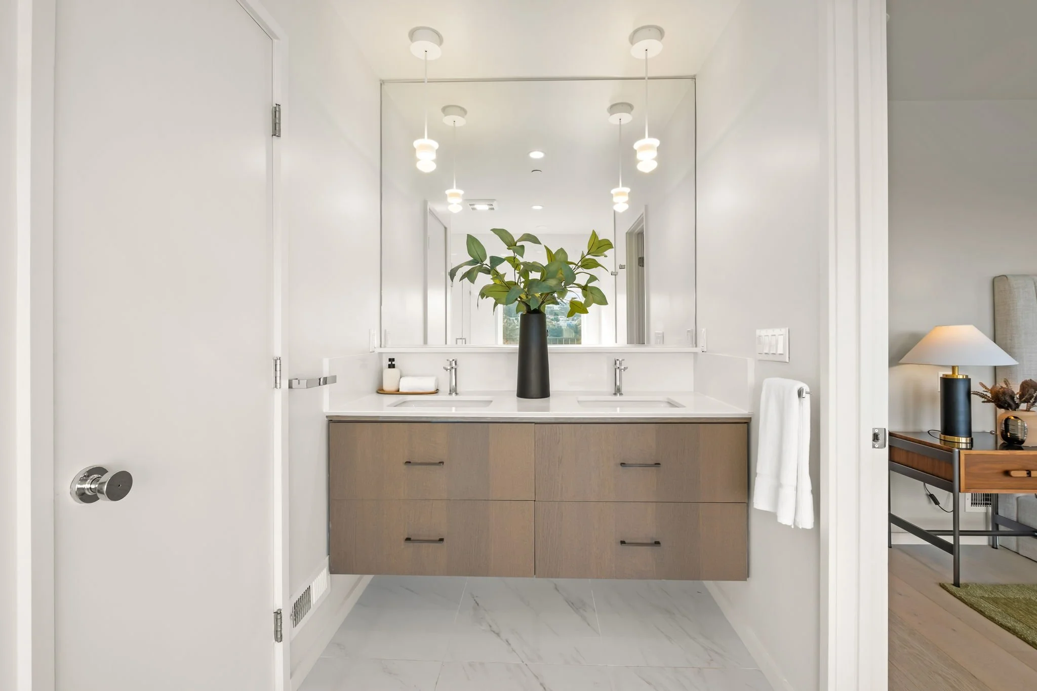 Modern bathroom with a double sink vanity, large mirror, and green plant in a black vase, with hanging pendant lights above.