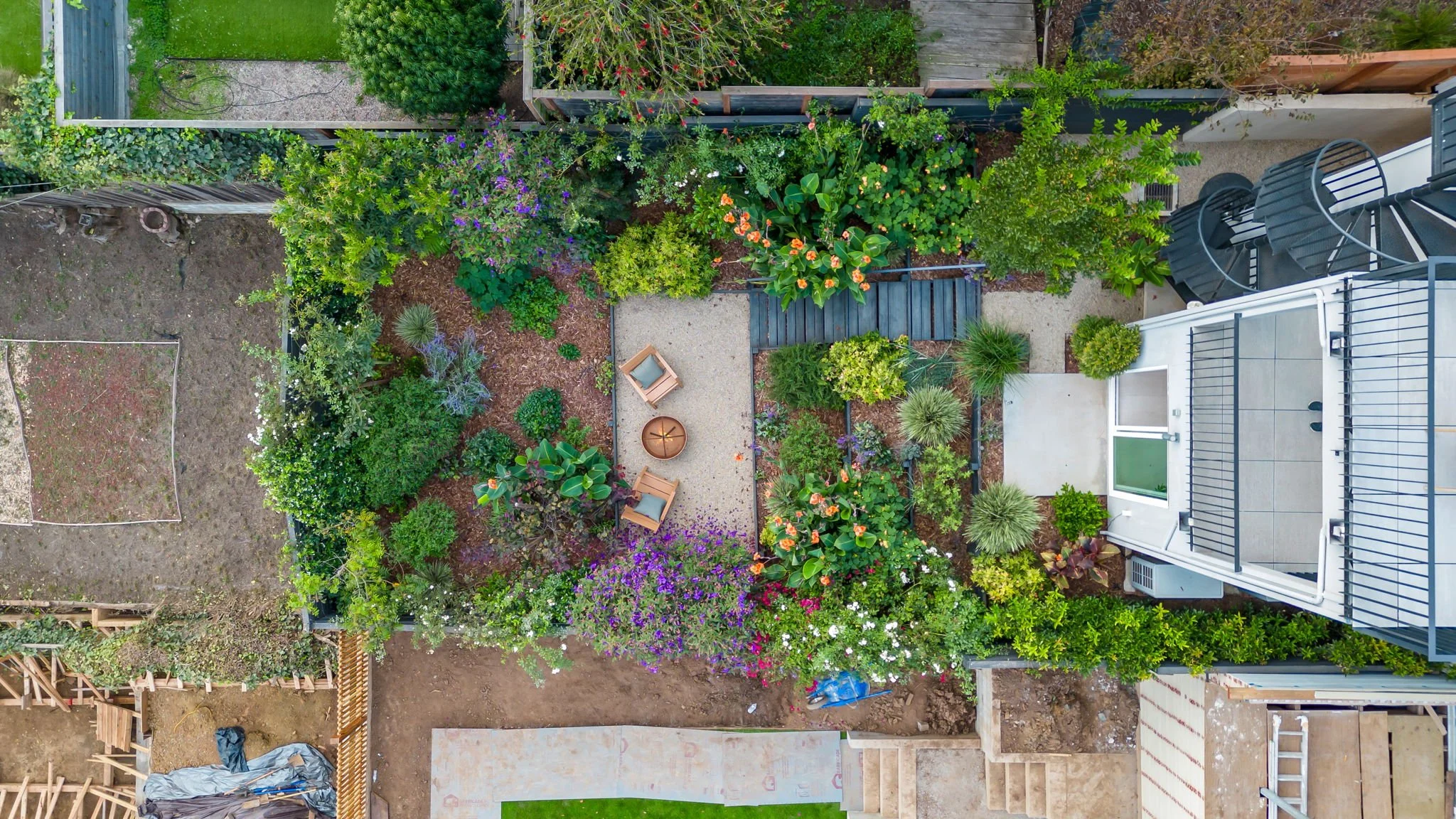 An aerial view of a backyard garden with lush green trees and bushes, colorful flowers, a small patio area with two chairs and a fire pit, adjacent to a house with a balcony and sliding glass door, with some areas under construction.