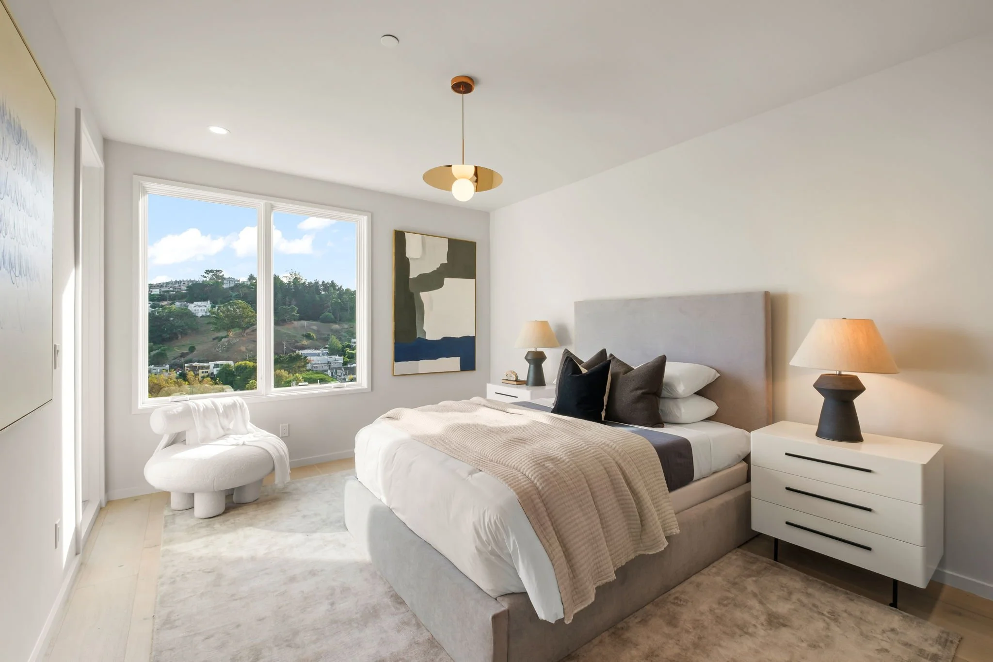 Modern bedroom with a large bed, two nightstands with lamps, a window with a view of hills and houses, and abstract wall art.