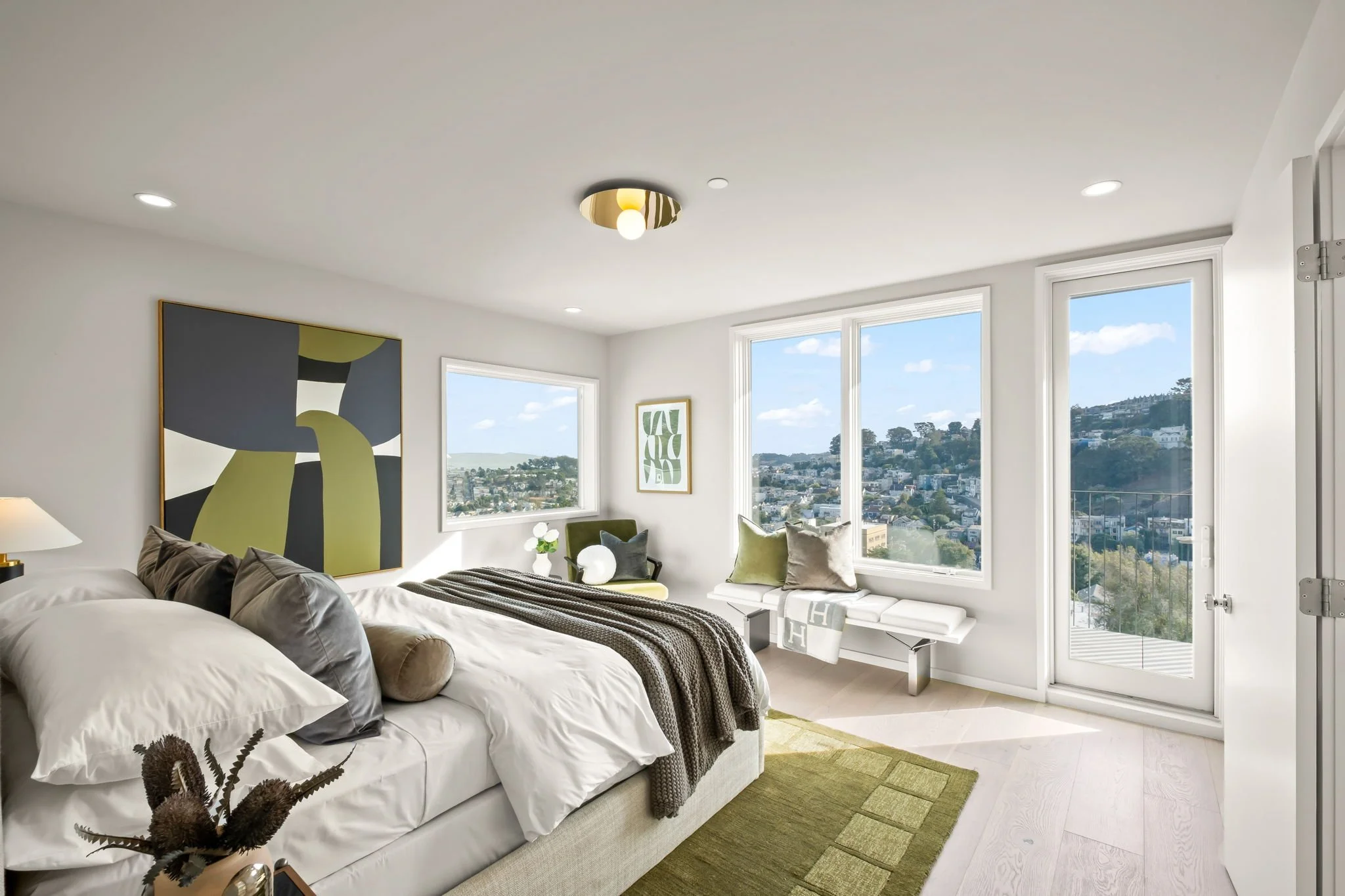 Bright modern bedroom with large windows overlooking a cityscape, featuring a bed with white and gray bedding, artwork on the wall, a bench with cushions, and a green rug.