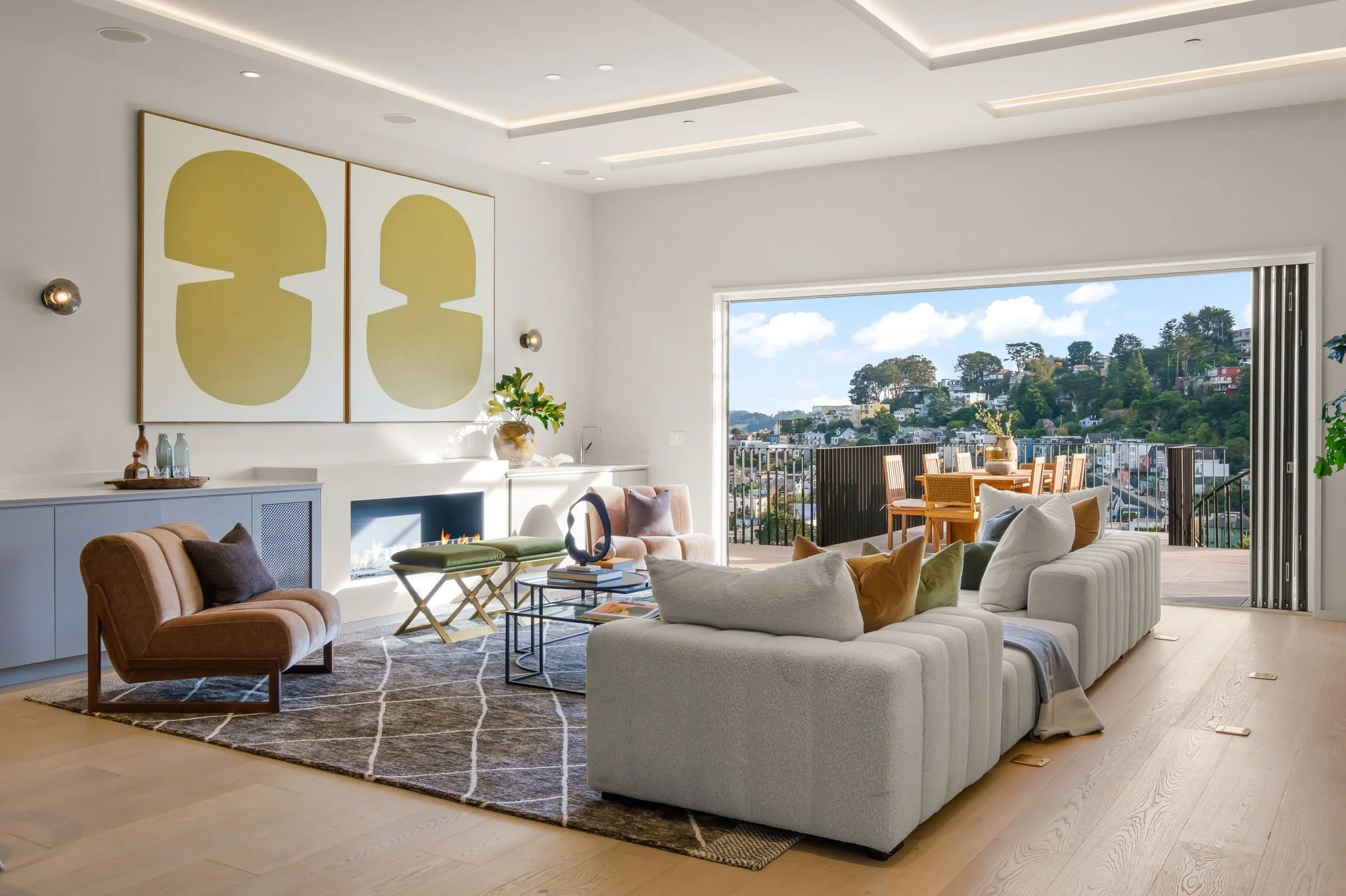 Modern living room with a white sectional sofa, brown armchair, and glass coffee table. Large sliding door open to an outdoor balcony with a view of a hillside neighborhood and blue sky.