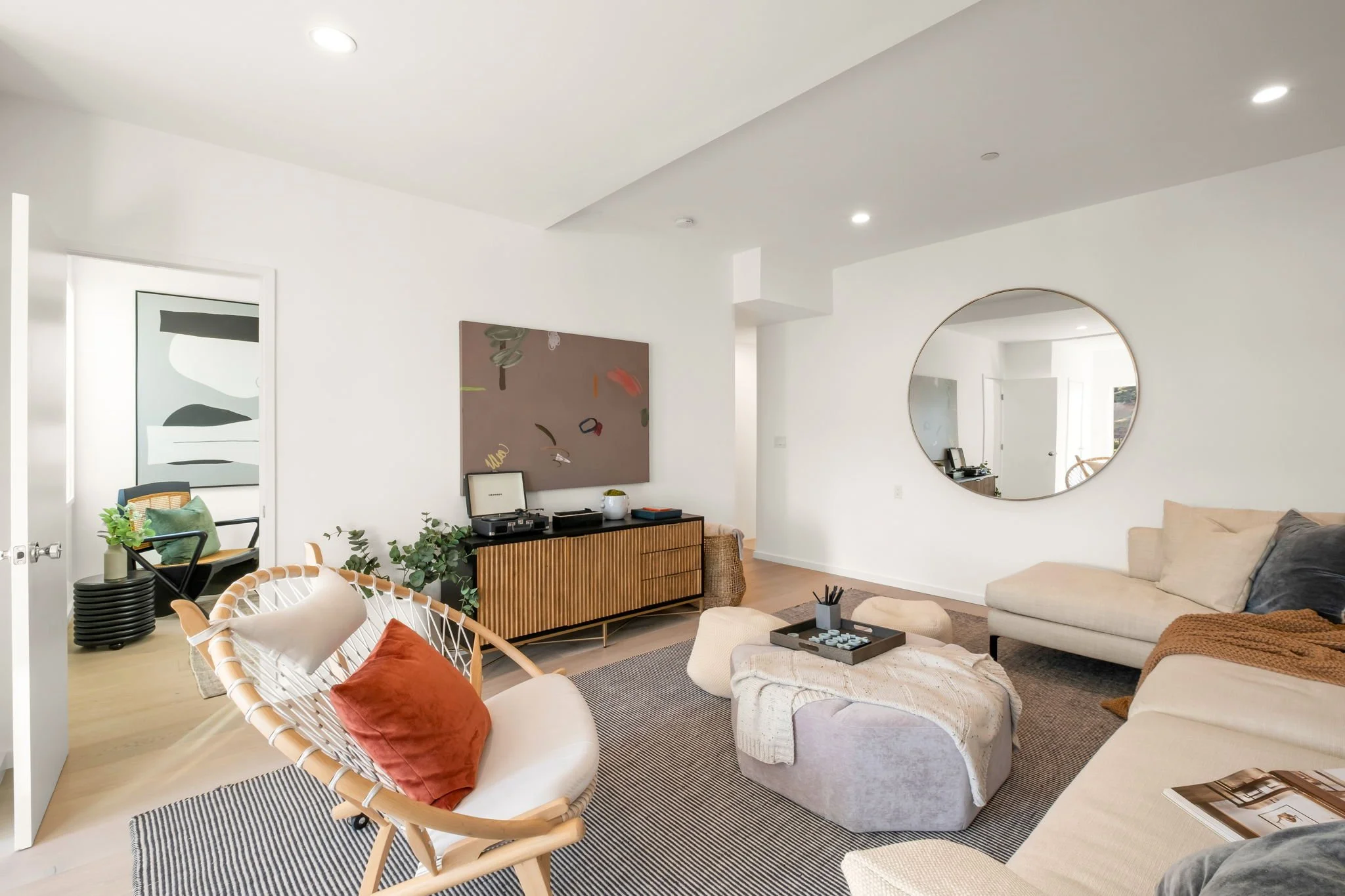 Modern living room with white walls and ceiling, featuring a large round mirror on the wall, beige sectional sofa with gray pillows, a wooden coffee table with black container holding pens, a cream woven chair with a rust orange pillow, a multicolore