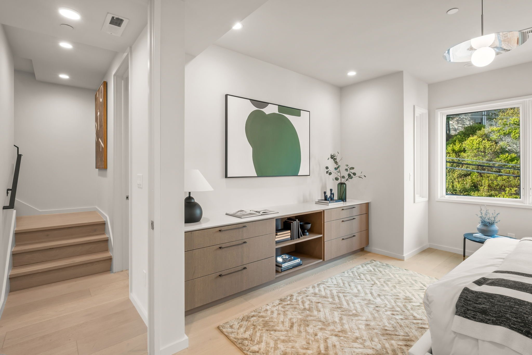 Bedroom with a wooden dresser, abstract green artwork on the wall, bed with black and white striped blanket, window showing green trees outside, lamp, and decorative objects on the dresser.