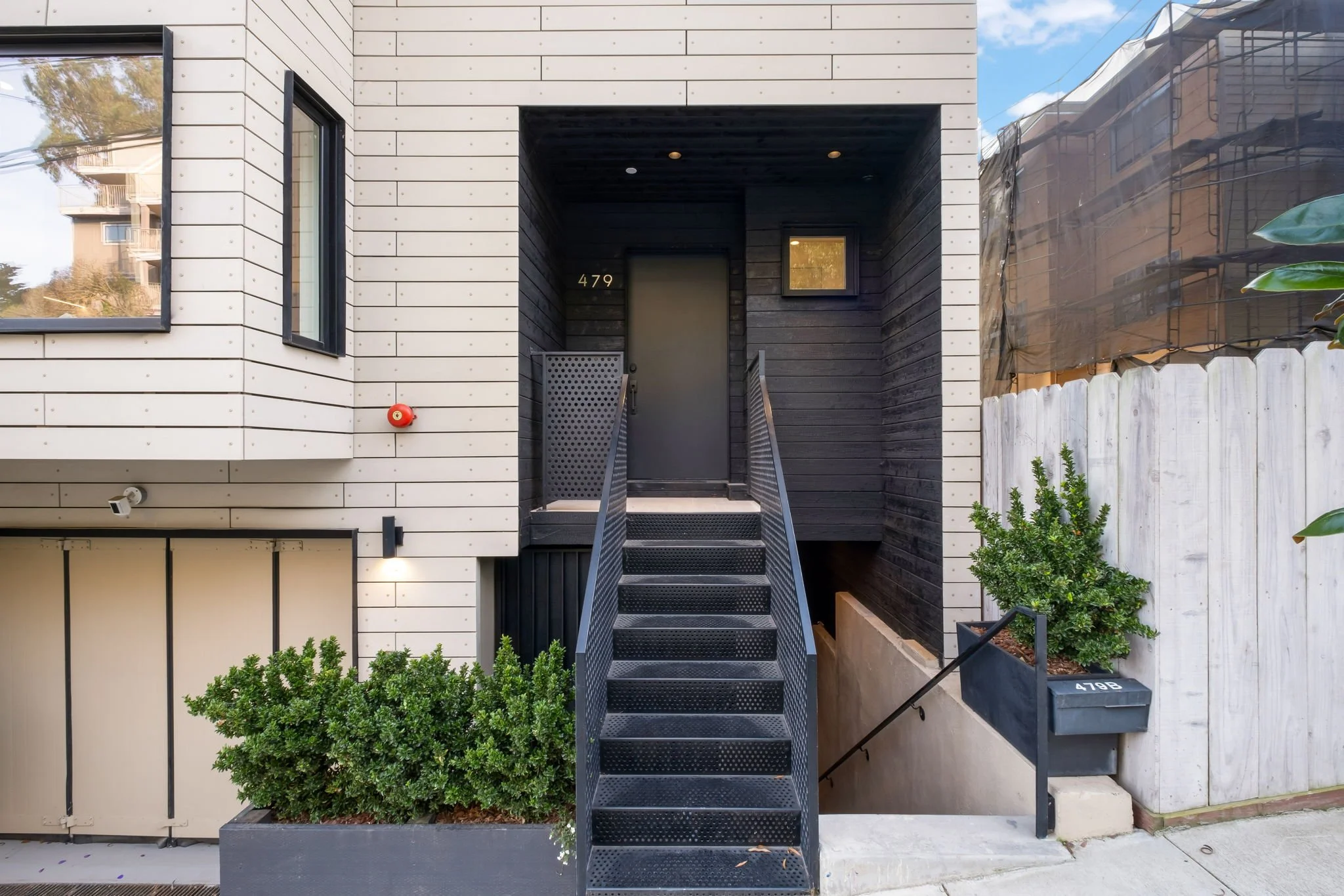 Front entrance of a modern house with a black front door, metal stairs leading up to the door, and a small window next to it. The house has light-colored siding with black accents and surrounding greenery.