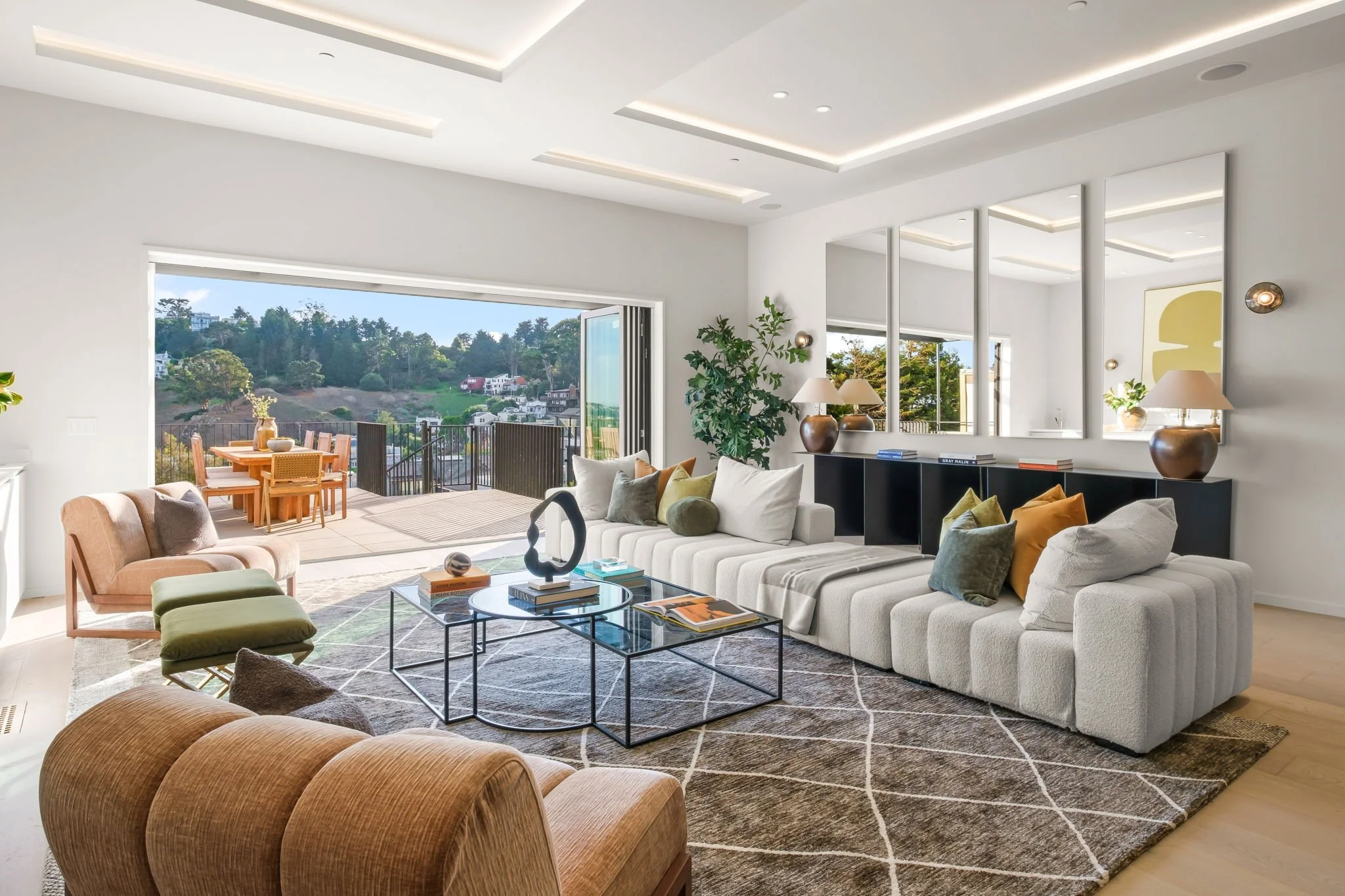 Modern living room with large window opening to an outdoor patio and scenic hillside view, featuring a beige sofa with colorful cushions, a glass coffee table with books and decorative items, a plush peach-colored armchair, a green ottoman, a black console with lamps and mirrors, and framed art on the wall.
