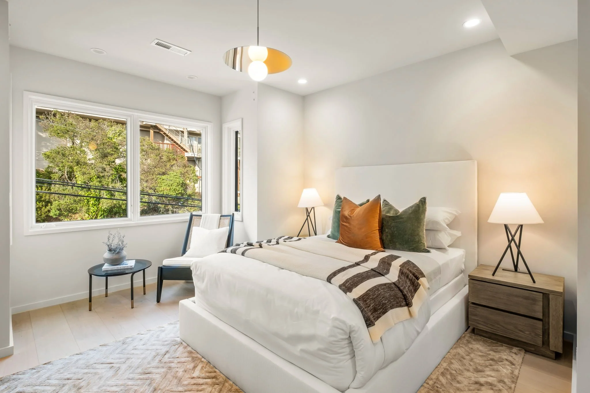 Modern bedroom with a white bed, colorful pillows, wooden nightstand, table lamps, large window with greenery outside, and a sitting chair near the window.