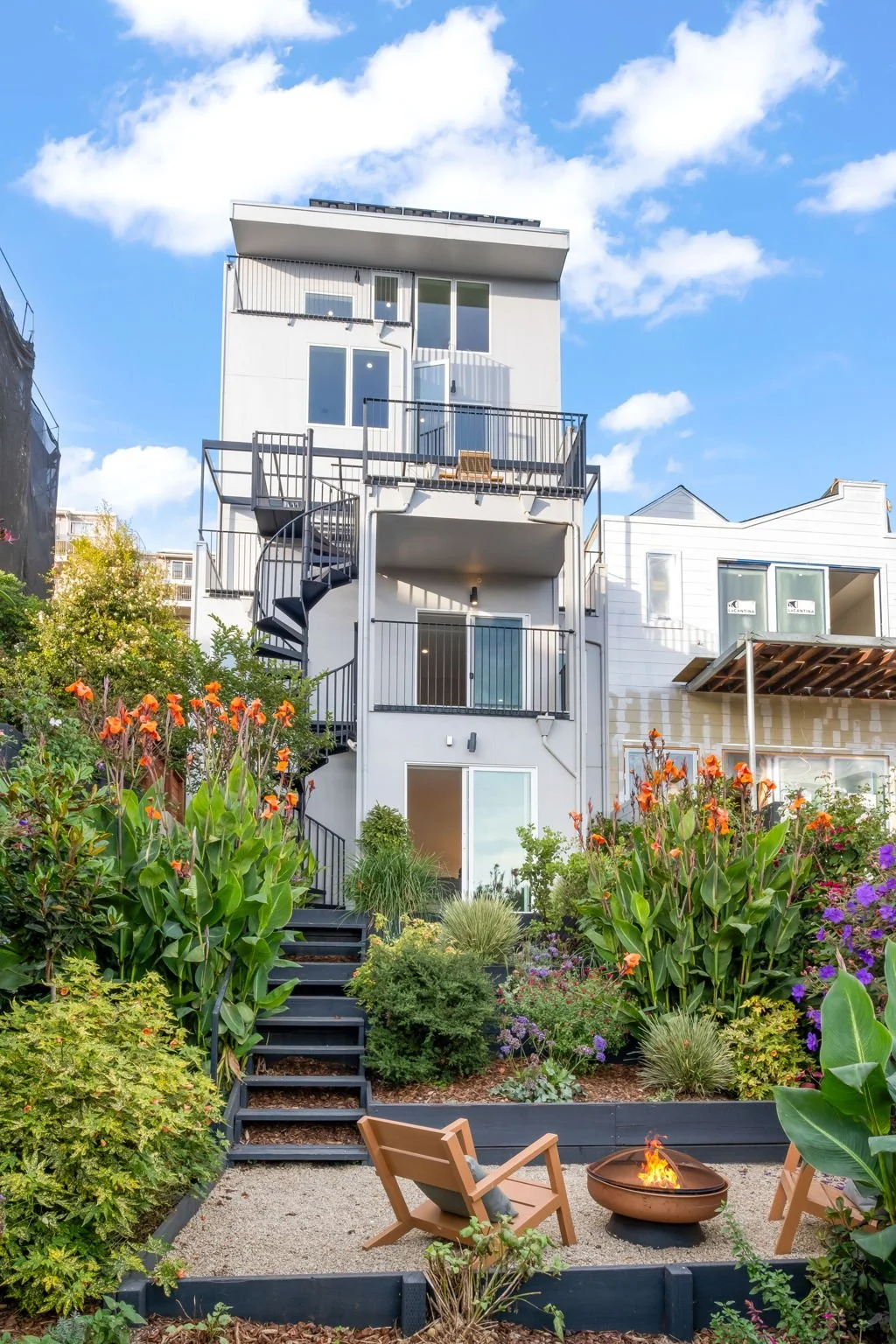 Multi-story white modern house with outdoor spiral staircase, surrounded by lush garden with orange and purple flowers, small fire pit, and two wooden chairs, under a bright blue sky with clouds.