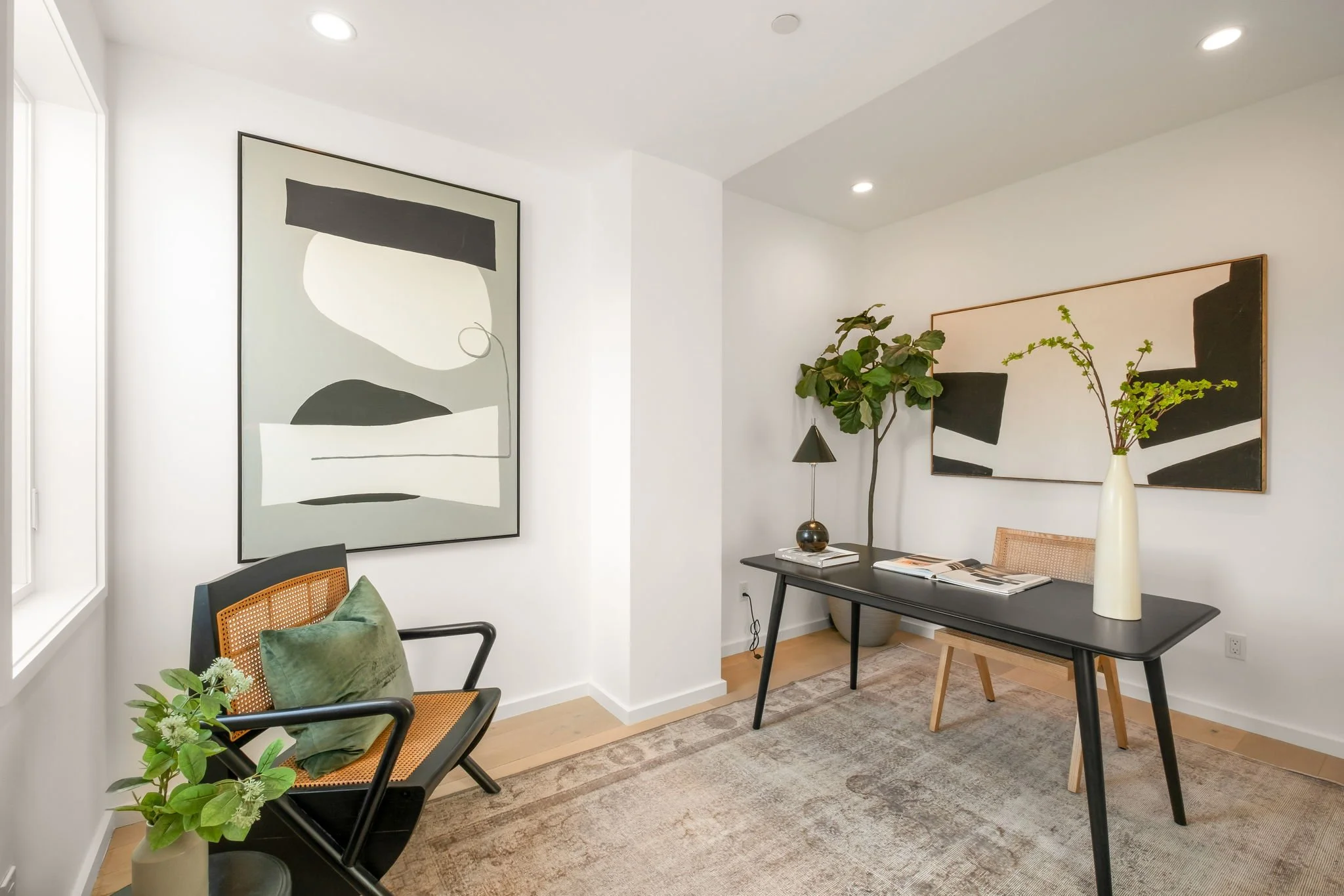 Modern home office with a black desk, a wooden chair with woven backrest, a large green plant, abstract black and white artwork, and a beige area rug.