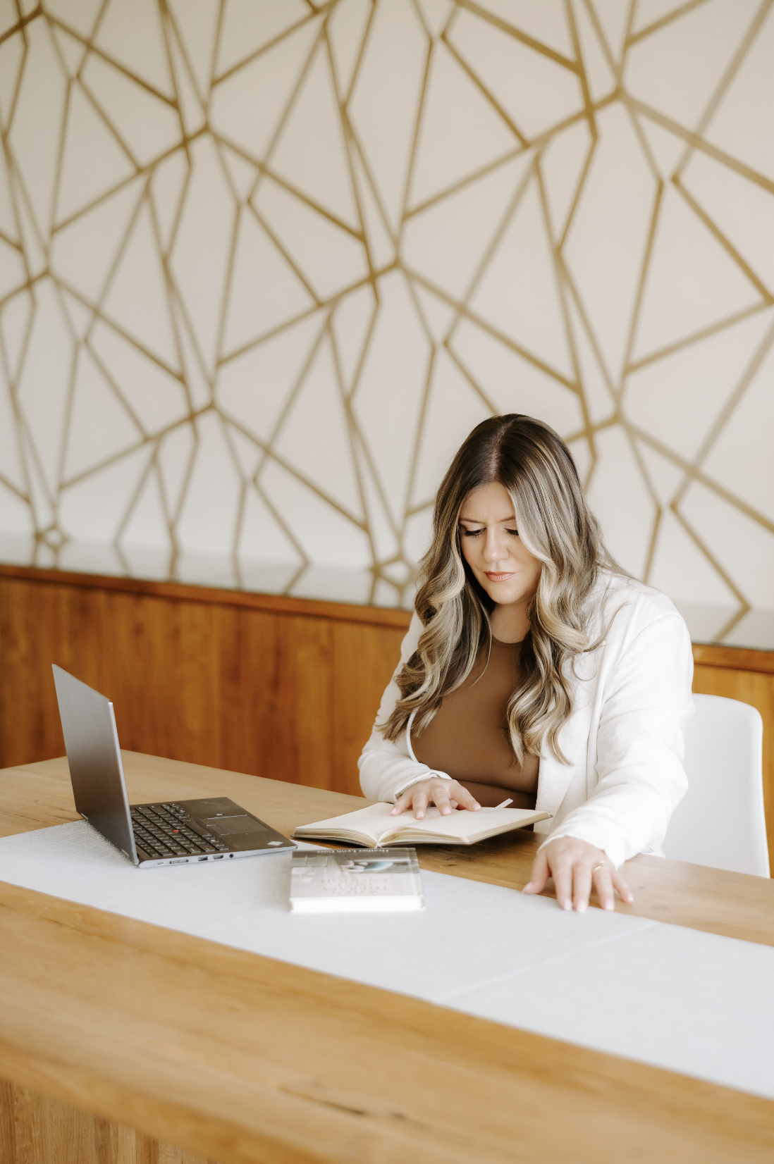 Heather Gorr, LPC-S, trauma-informed therapist and clinical supervisor based in Austin, Texas | Woman with dark curly hair, wearing a white suit with a brown shirt, sitting at a table reading a book with a laptop open