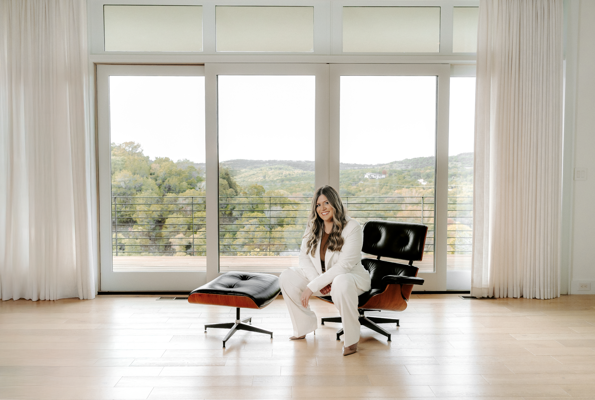 A woman in a white suit sitting on a black and wood lounge chair in a bright room with large glass door and window overlooking a green landscape.